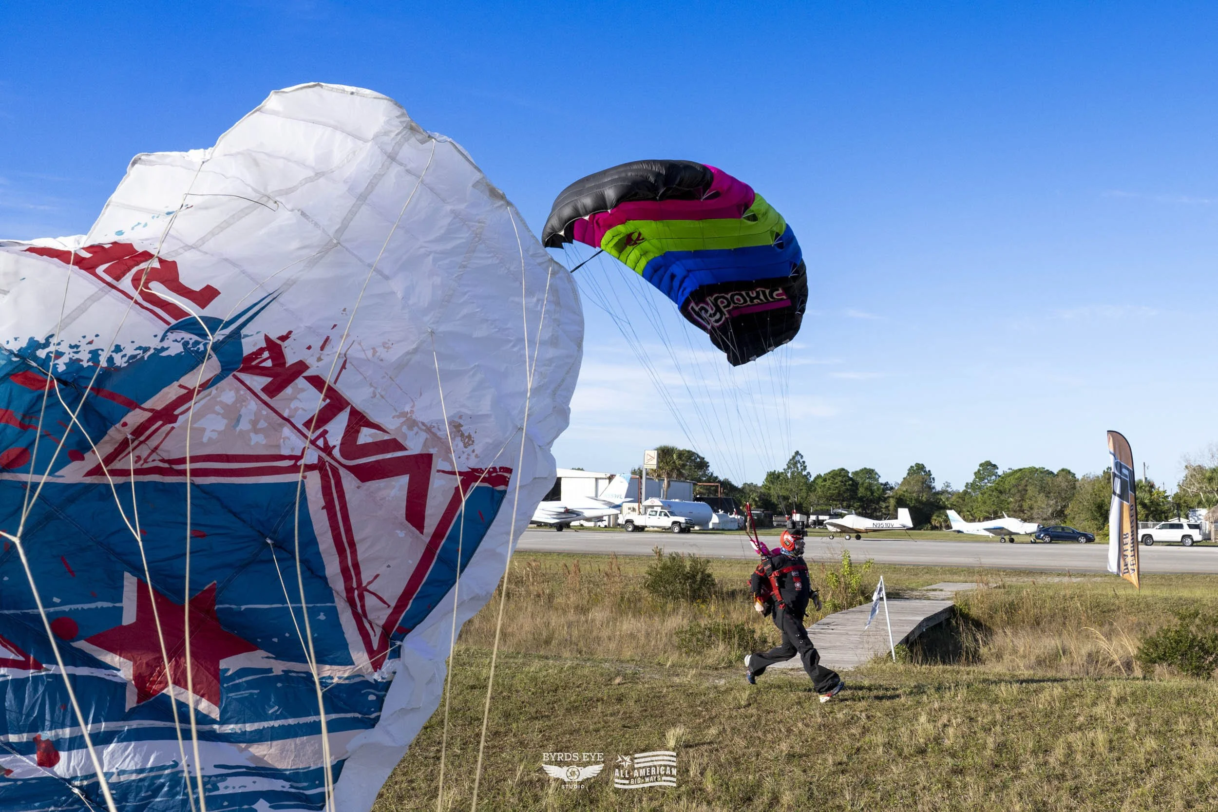 A person jogging near a grassy area with parachutes and small planes in the background at an airfield, under a clear blue sky.