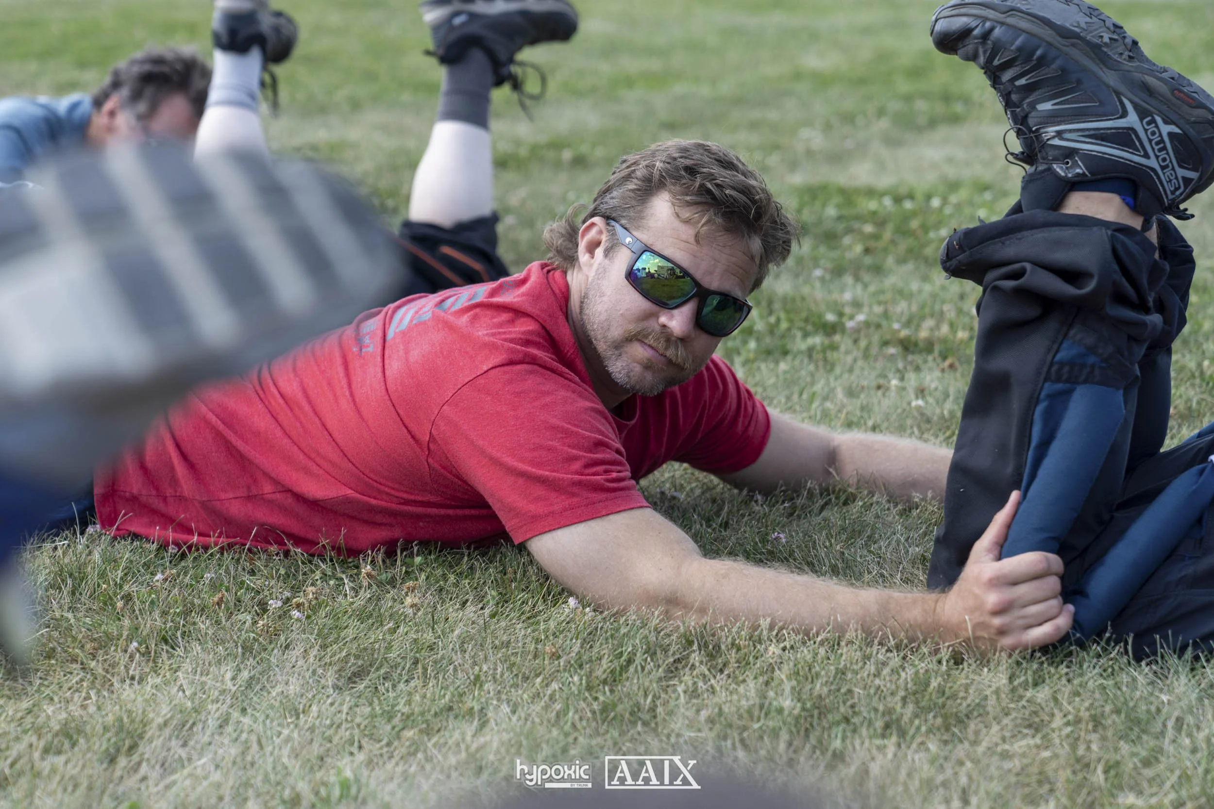 A man in a red shirt and sunglasses lying on grass next to a backpack, with another person in the background also lying on the grass.