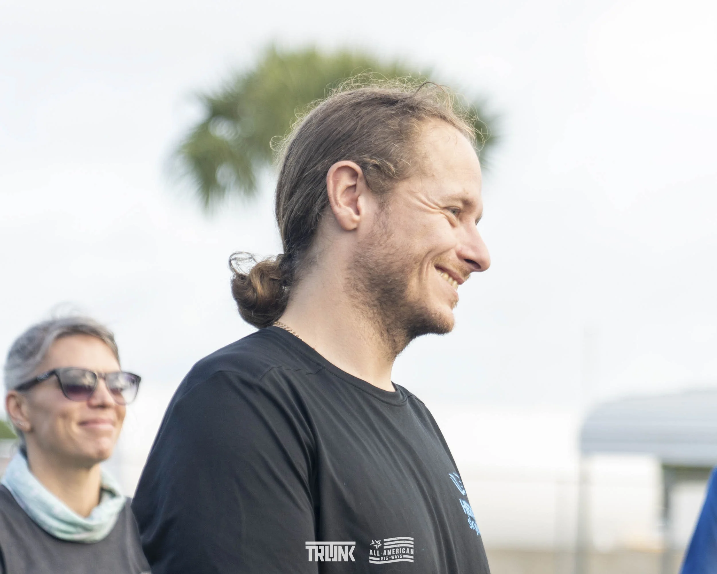 A man with long hair tied back, smiling while looking down, standing outdoors. A woman with glasses and short hair, wearing a neck gaiter, stands behind him.
