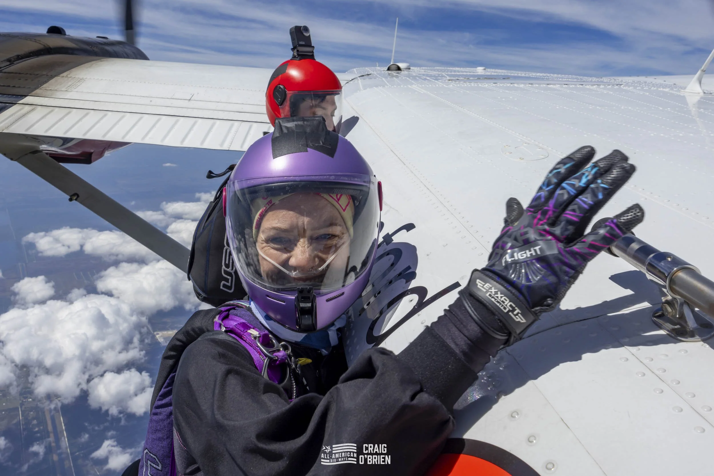Two skydivers on an airplane, one in a purple helmet and the other in a red helmet, taking a selfie before a jump. The woman in the foreground is smiling and waving, with clouds and blue sky in the background.
