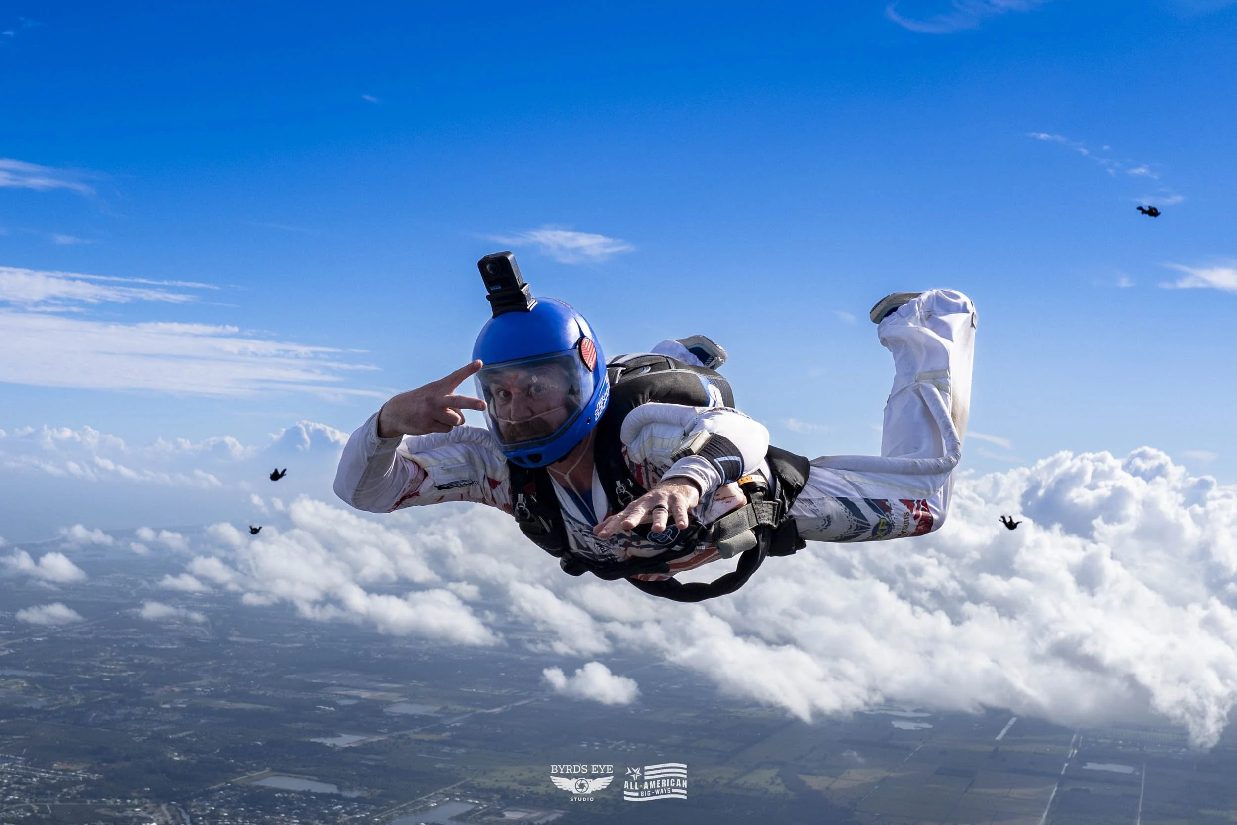 A man skydiving mid-air wearing a blue helmet with a camera, white jumpsuit, and a harness with a backpack, against a blue sky with clouds