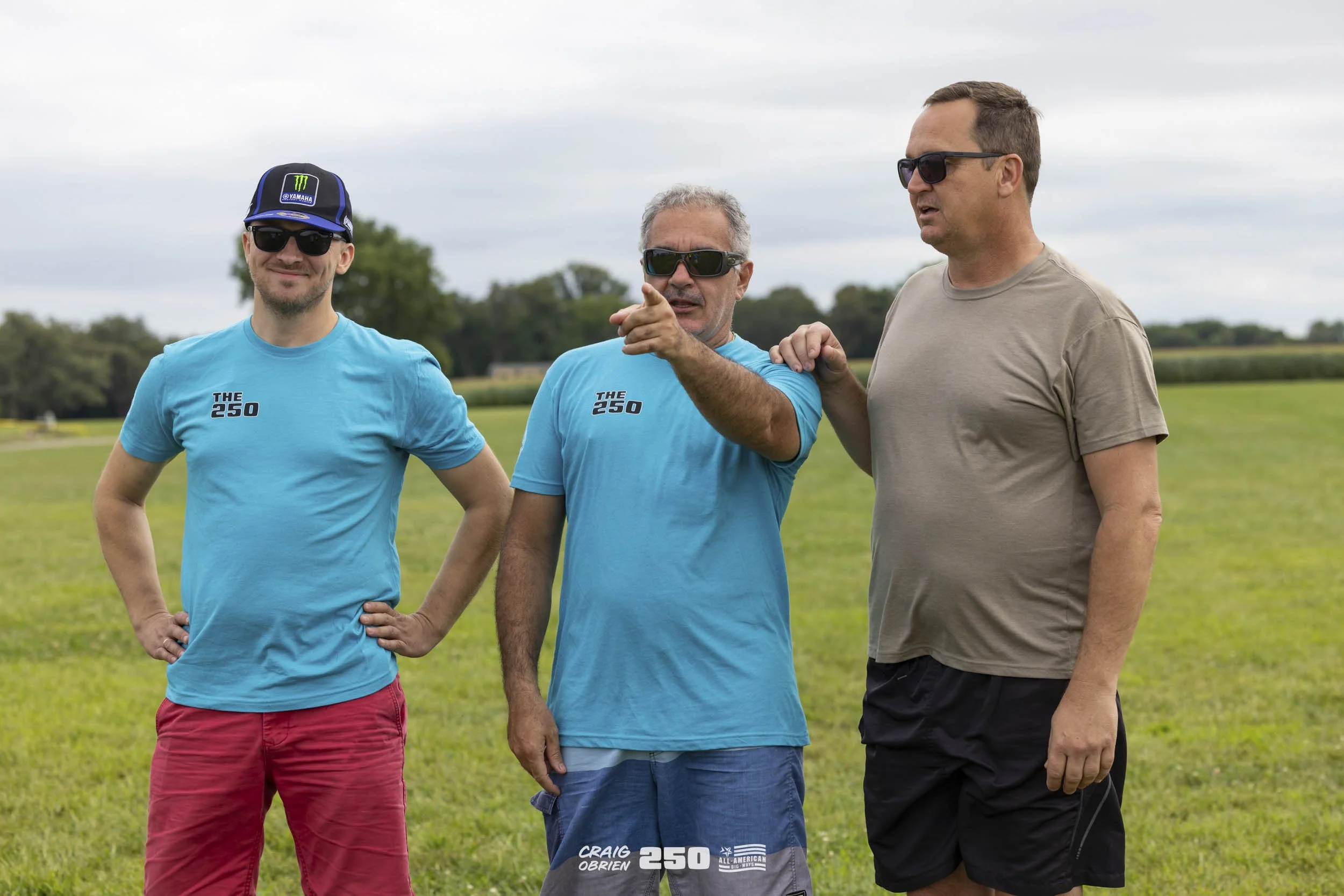 Three men standing on a grassy field, two wearing matching blue shirts with 'The 250' logo, sunglasses, and shorts, one in a beige shirt and black shorts, one man pointing and talking, overcast sky in the background.