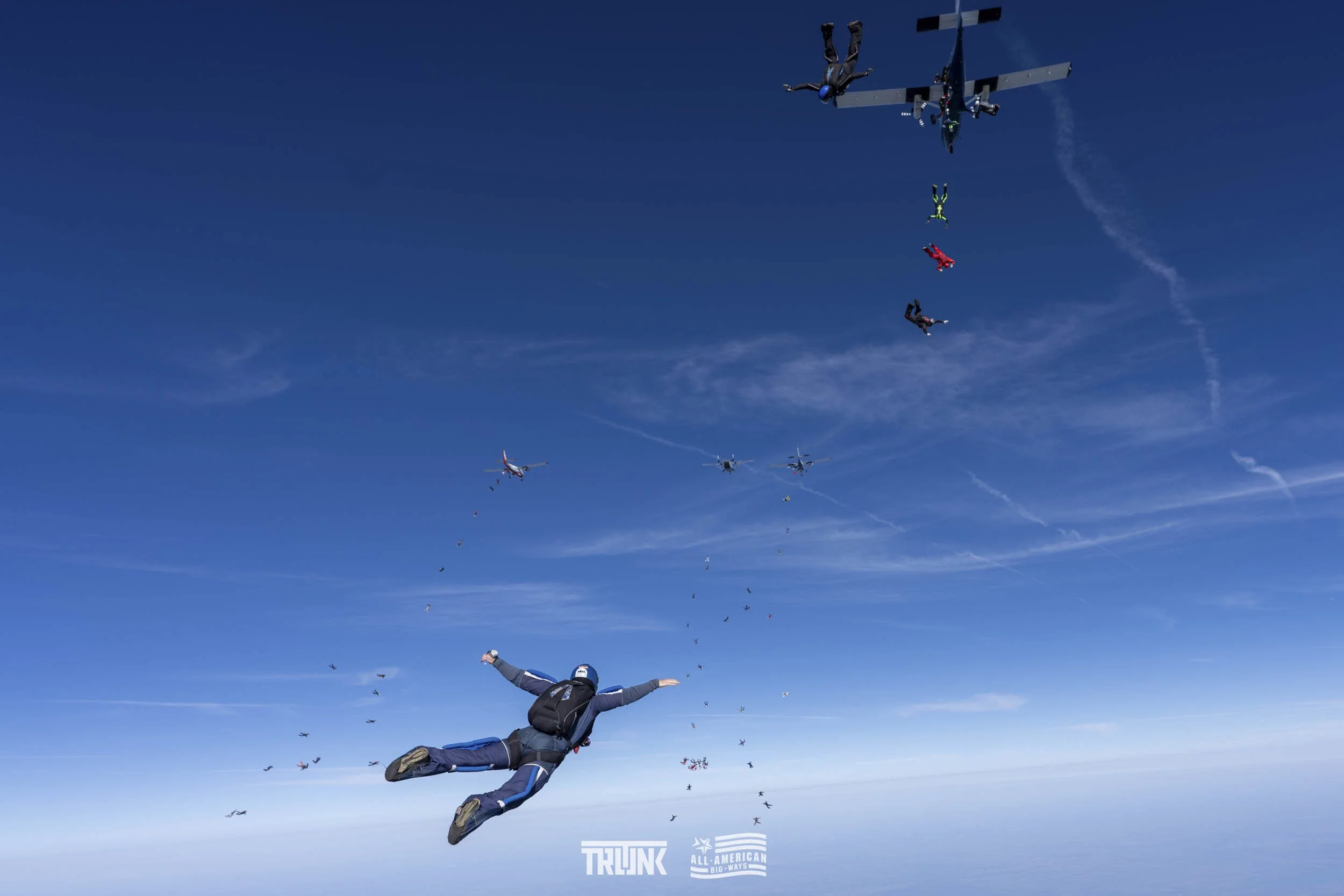 A skydiver in a jumpsuit falling through the sky with freefall posture, with several airplanes in the background, releasing more skydivers and parachutes, under a blue sky with wispy clouds.