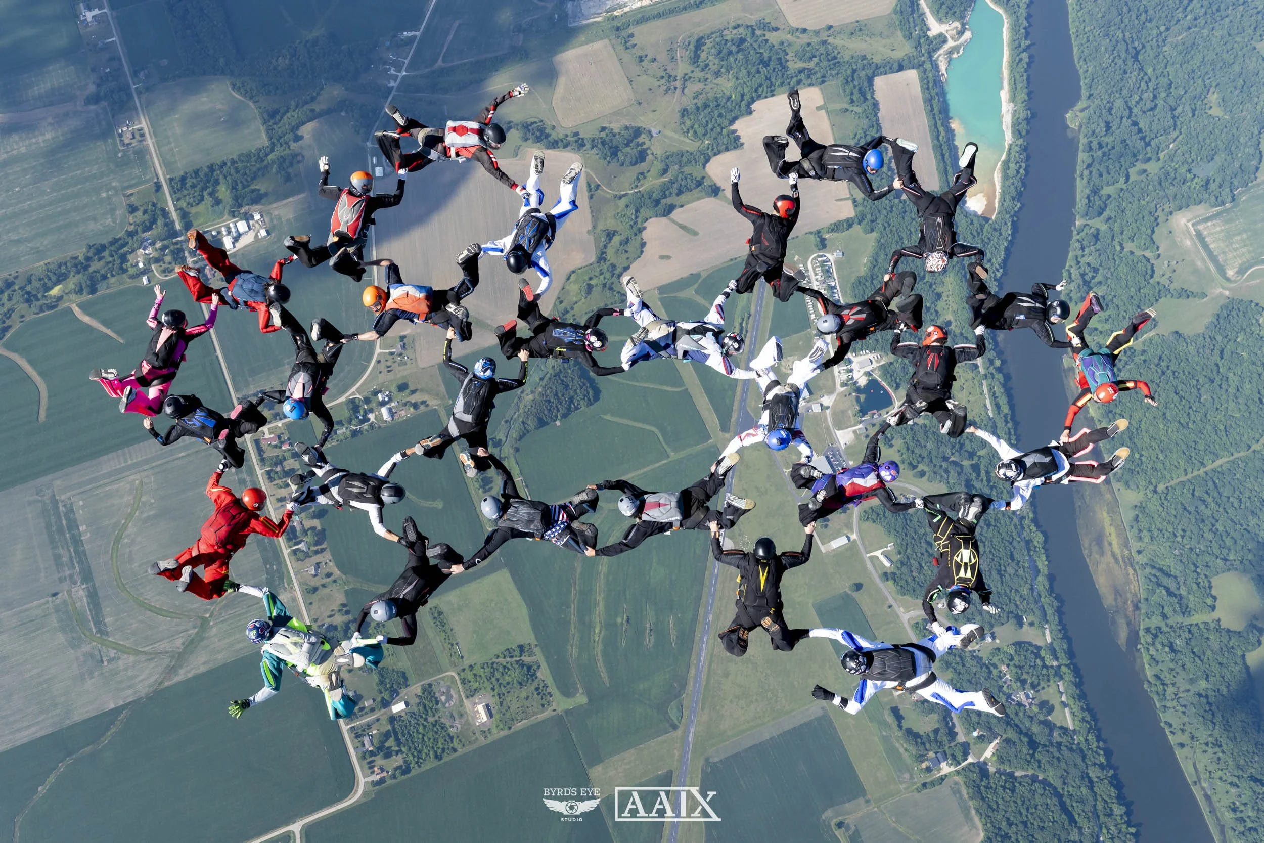 Group of skydivers in free fall holding hands in a circle over a rural landscape with fields, trees, a river, and a bridge.