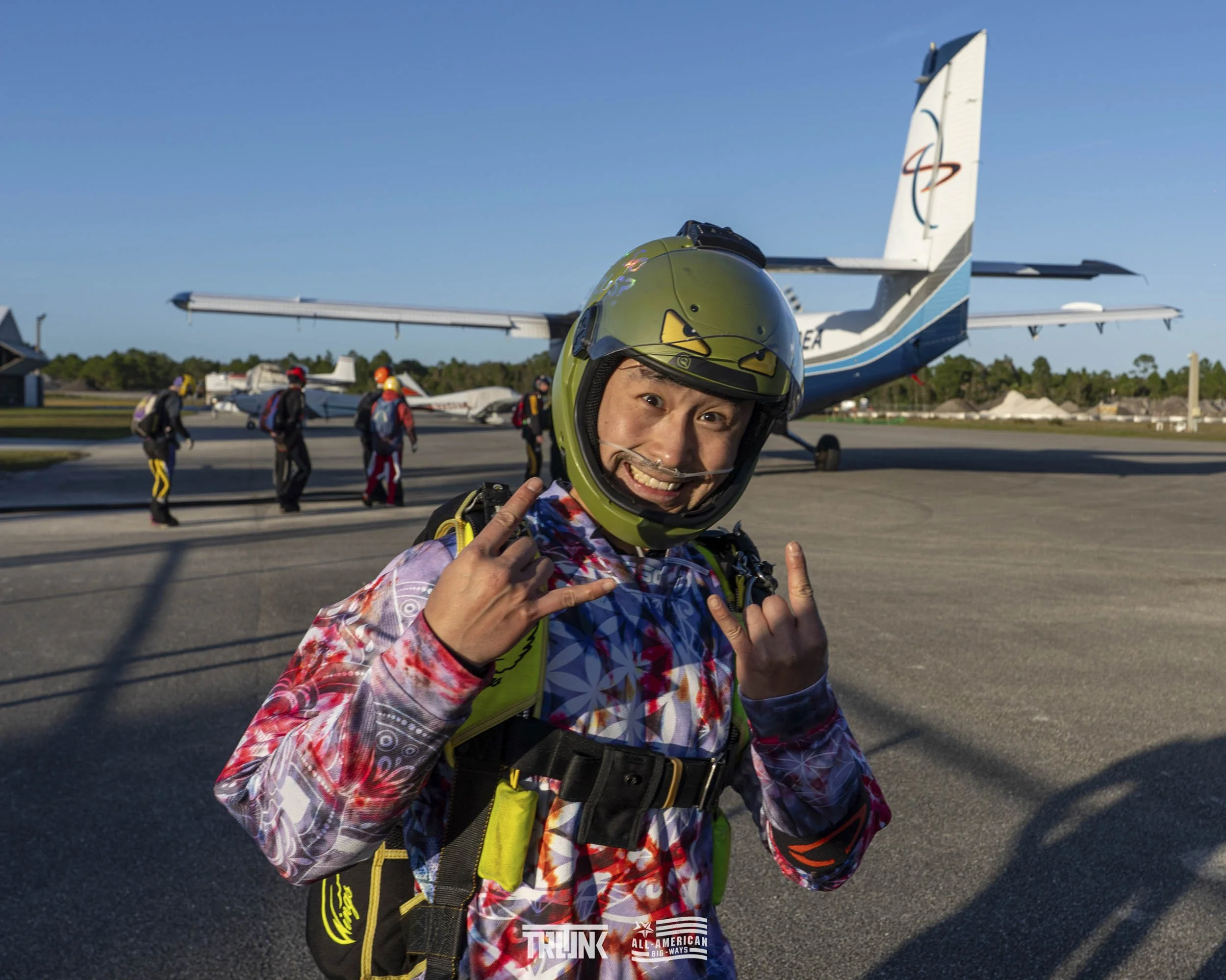 A skydiver wearing a helmet and jumpsuit making a rock and roll hand gesture on the tarmac with a plane and other skydivers in the background.