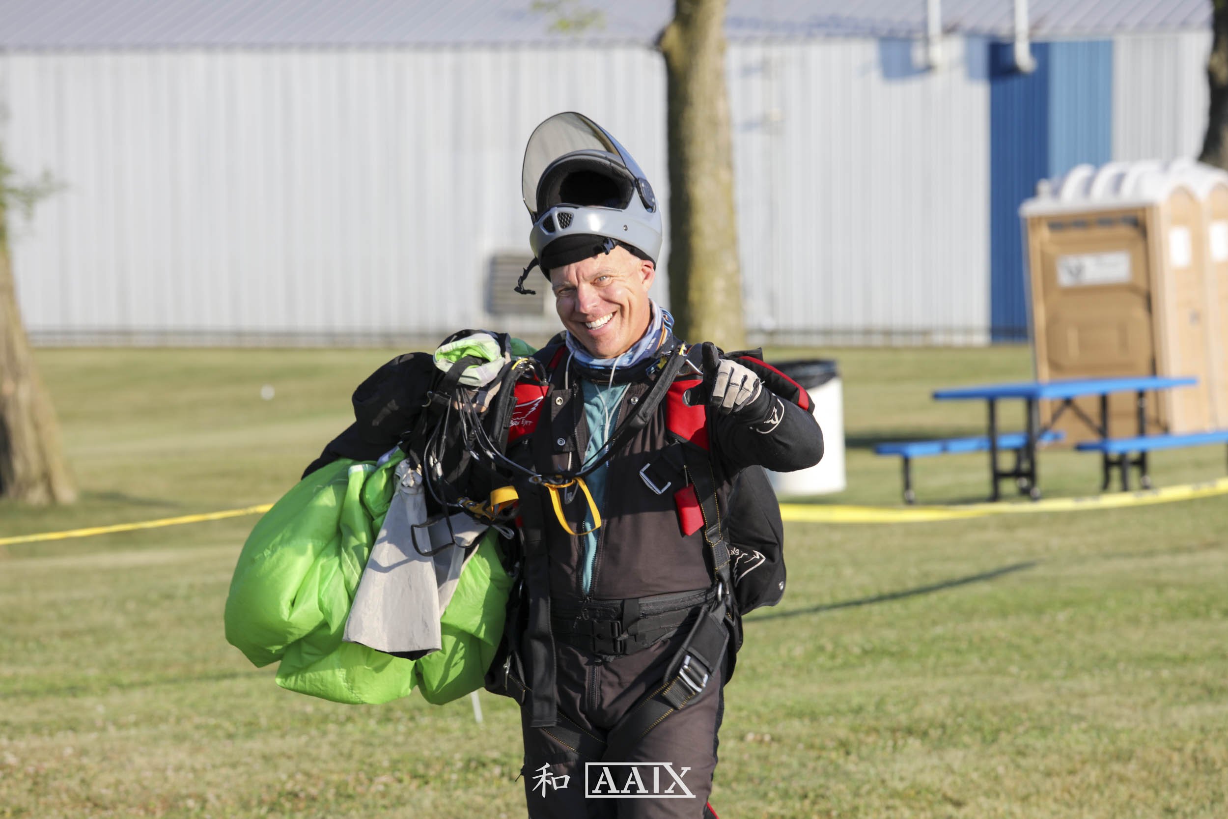 A man in skydiving gear smiling and pointing at the camera, with a green parachute on his shoulder, on a grassy field near a building, with a blue picnic table and portable toilets in the background.