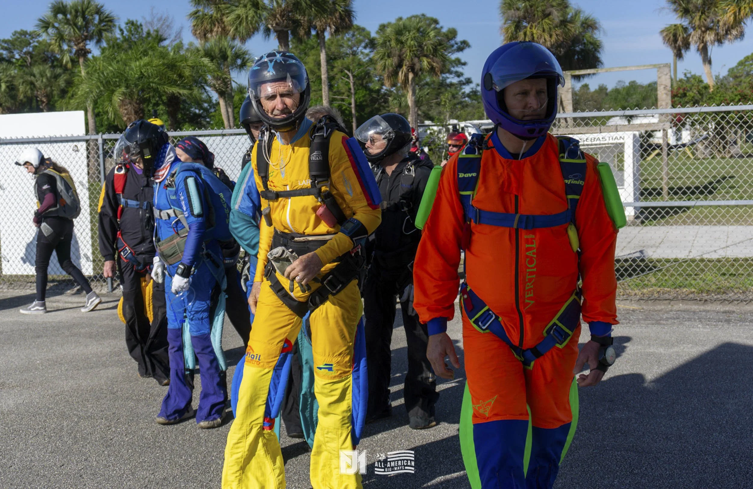 A group of skydivers in colorful jumpsuits and helmets standing outdoors on a paved area near a chain-link fence with palm trees in the background.