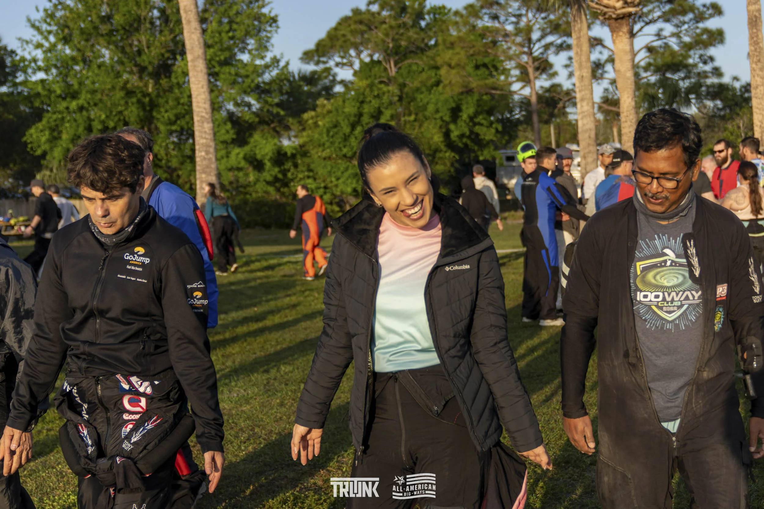 People walking outdoors on grassy area with trees in background, some wearing casual and athletic clothing, during sunny weather.