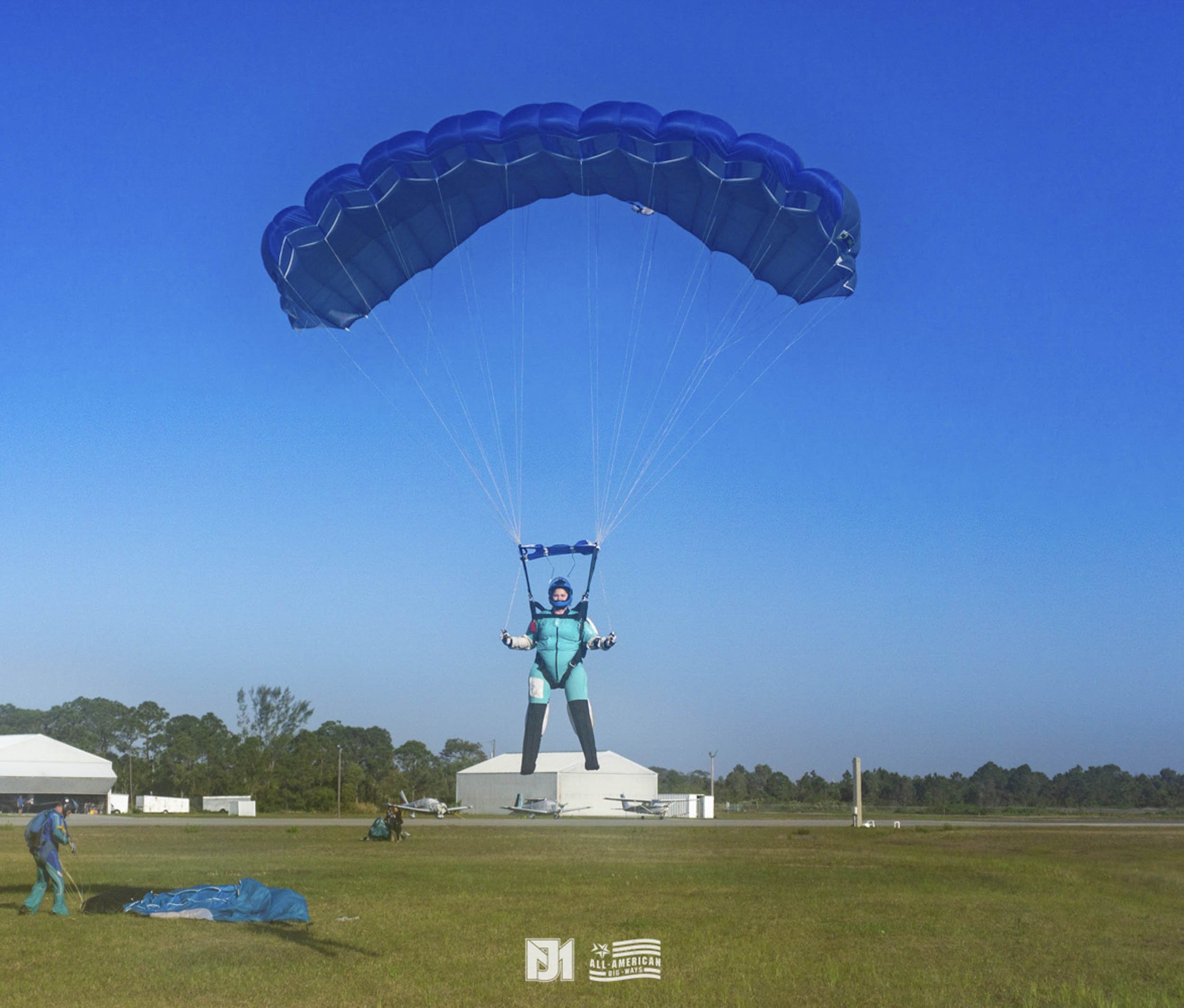 Person in a blue jumpsuit landing with a parachute on the ground at an airfield, with some structures and trees in the background.