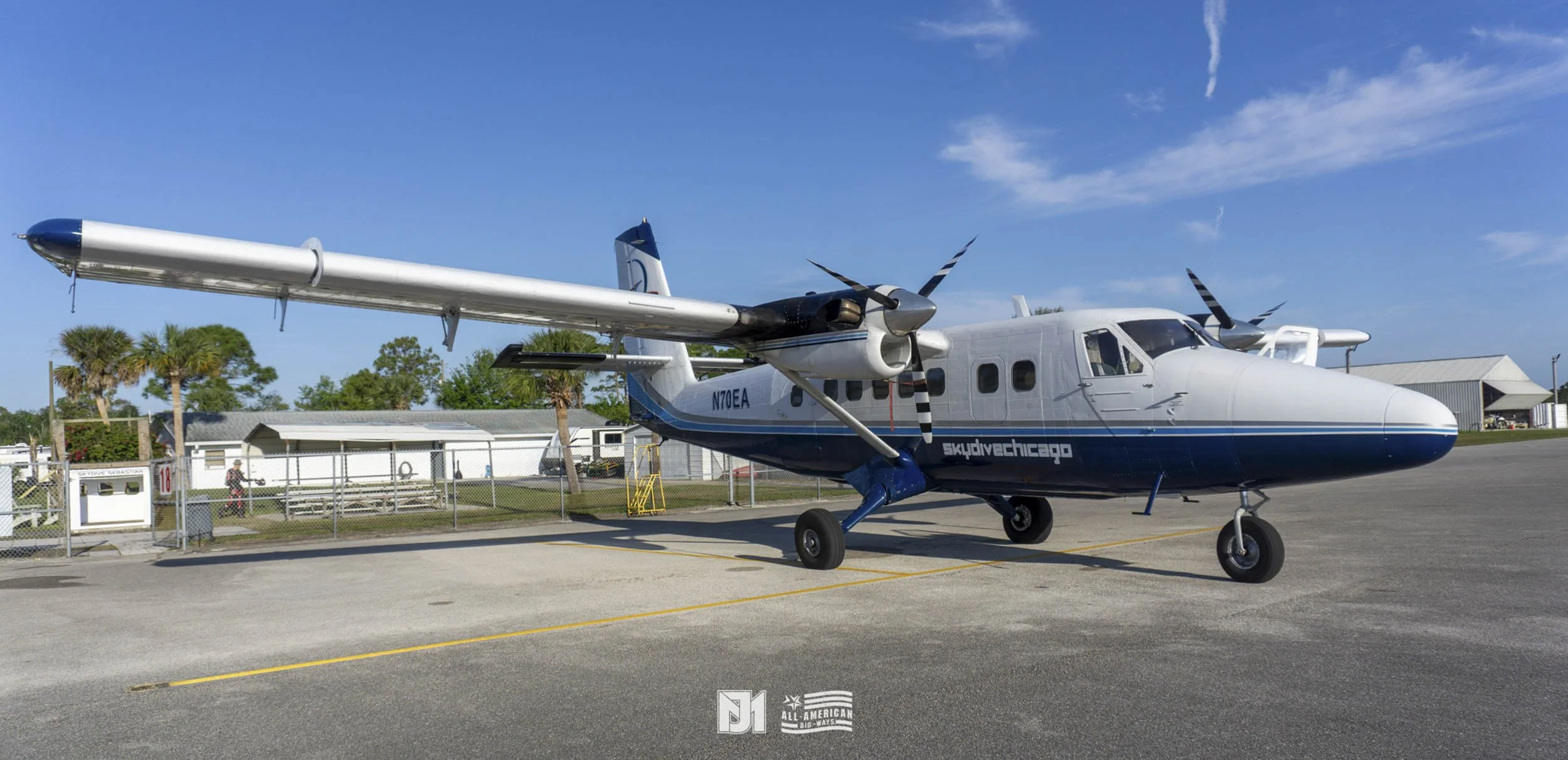 Small twin-engine airplane parked on tarmac, with buildings, trees, and a blue sky in the background.