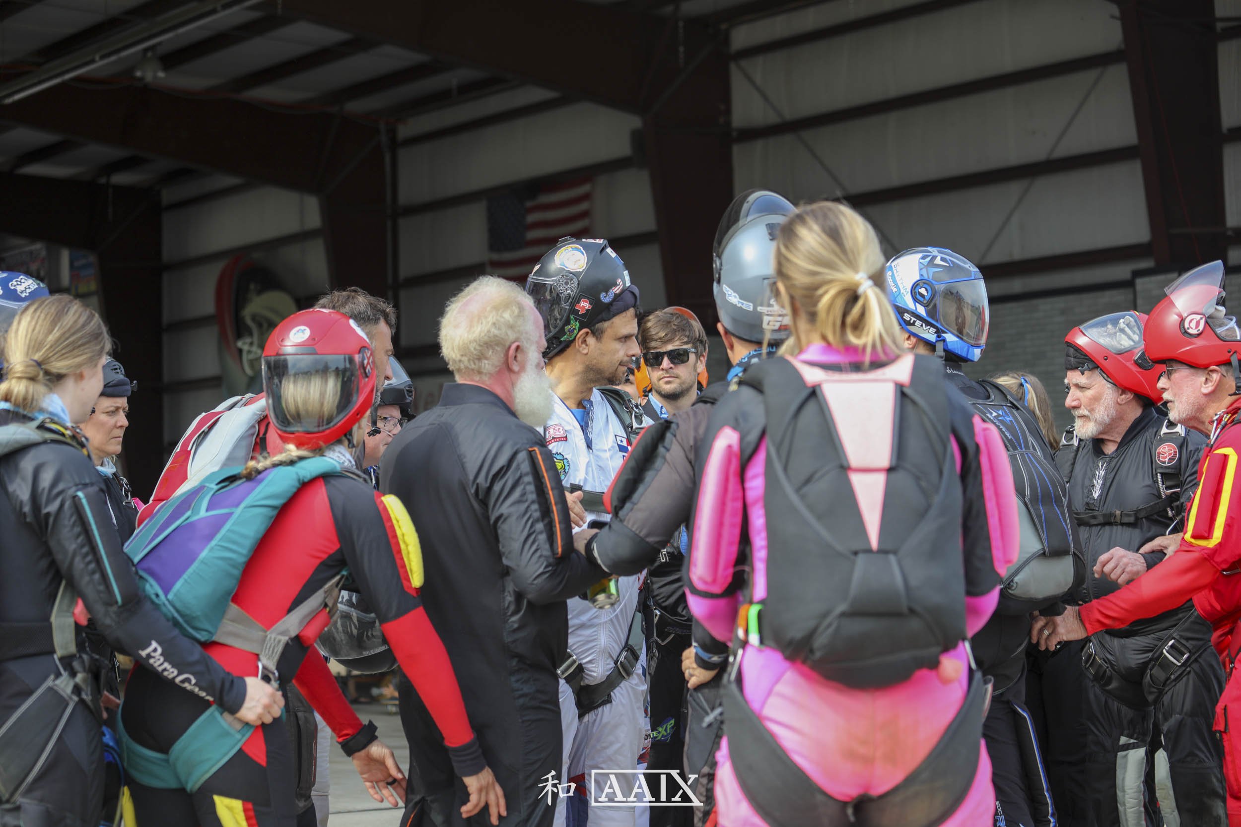 Group of people gathered inside a hangar, some wearing motorcycle helmets and gear, engaged in conversation, with an American flag hanging in the background.