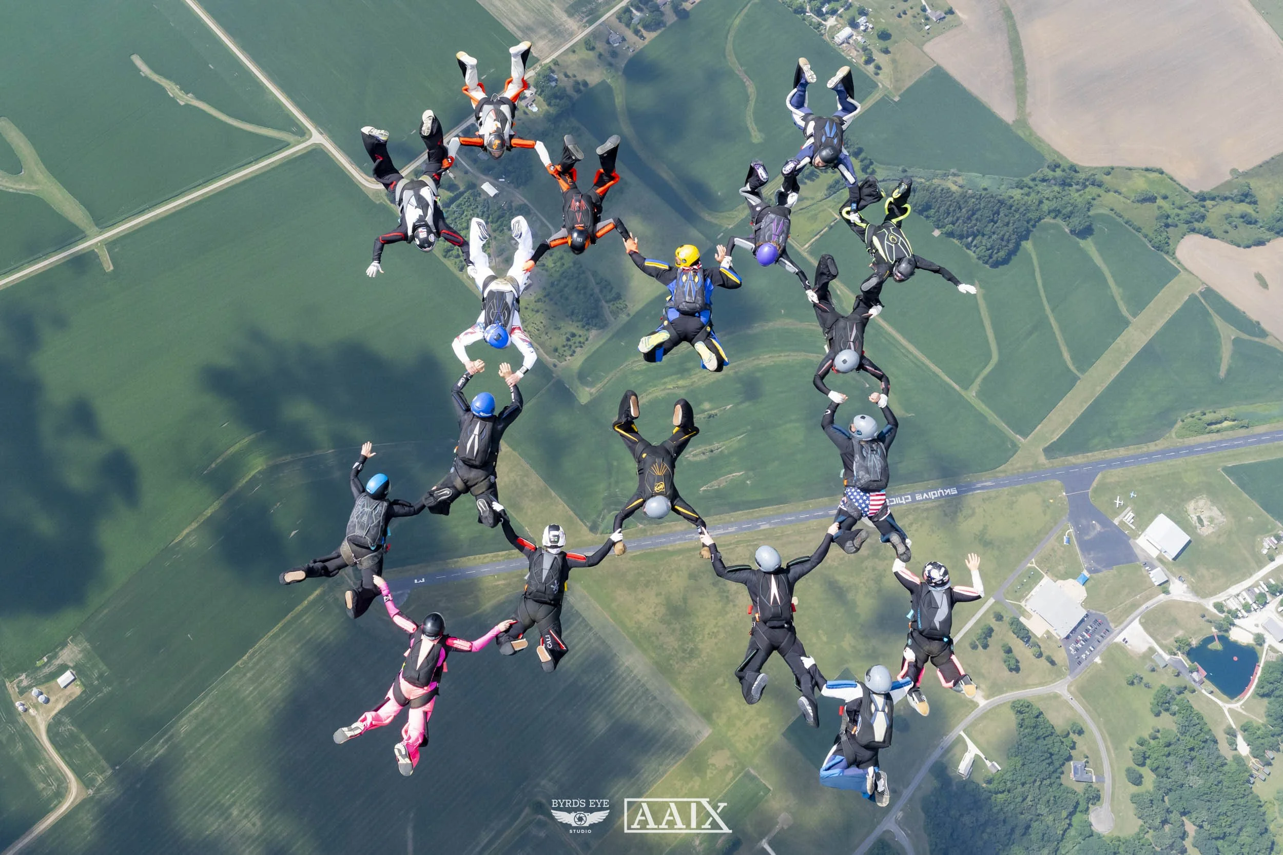 A group of skydivers forming a heart shape while free-falling over a rural landscape.