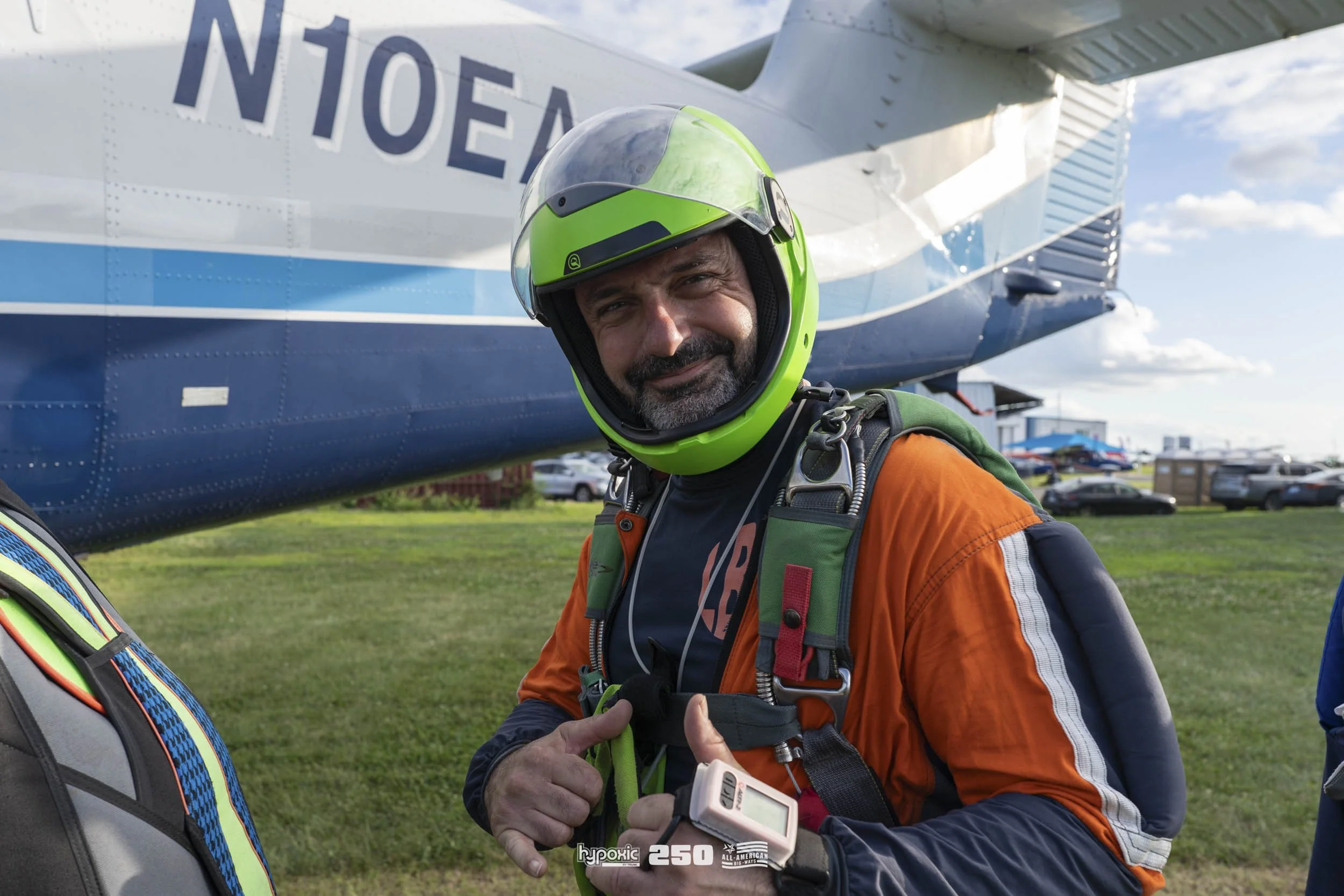 A man in a bright green helmet, orange and gray jumpsuit, and harness, smiling and giving a thumbs-up in front of a blue and white airplane on a grassy field.