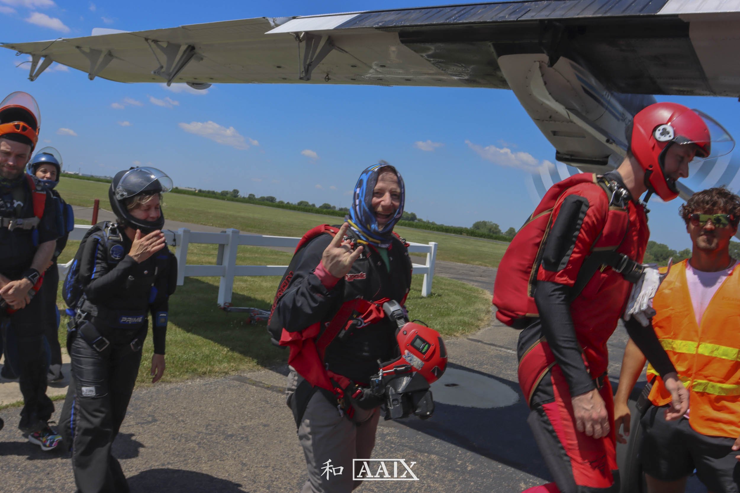 A group of skydivers in jumpsuits and helmets walking away from a plane on a sunny day with blue skies.