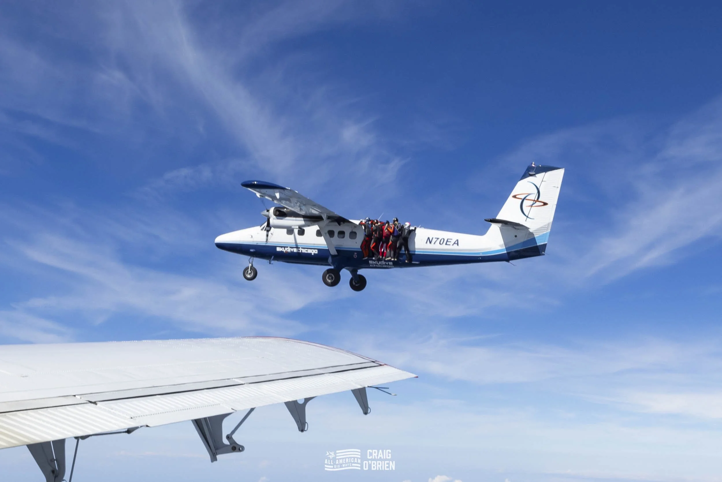 Scene of a small airplane in mid-flight with people parachuting out of it, against a blue sky with wispy clouds.