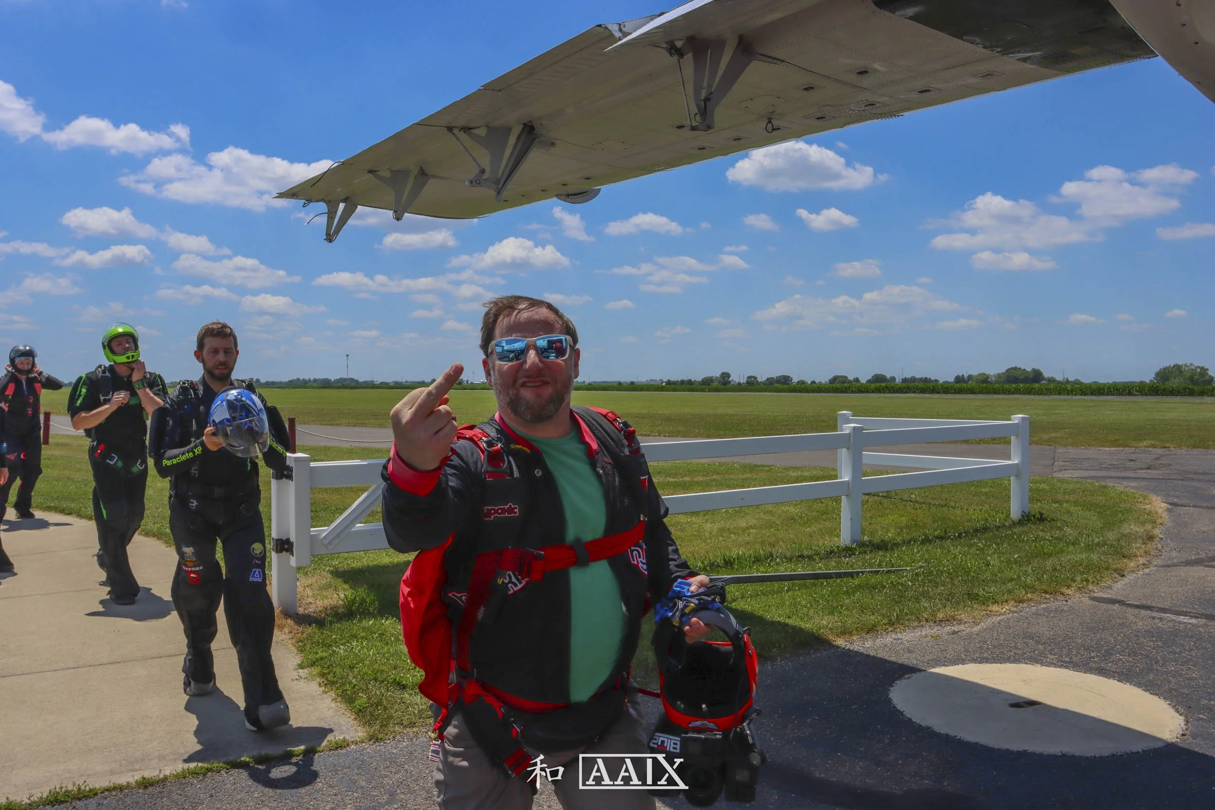 Group of skydivers walking away from a plane after landing, with one man in front showing middle finger, holding his helmet and wearing sunglasses, on a bright day with blue sky and clouds.