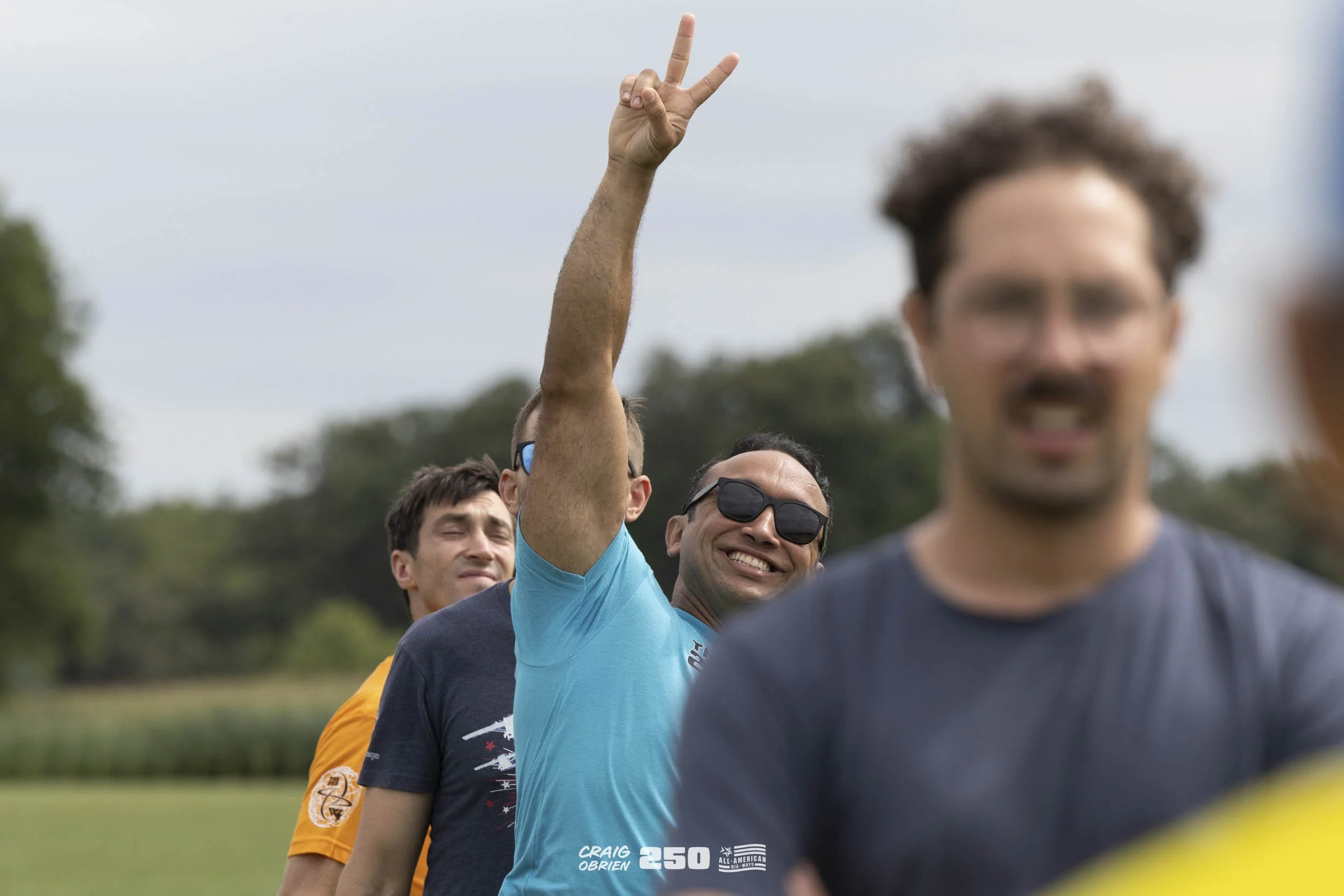 A group of men standing in a line outdoors, smiling and making peace signs with their fingers. The background features a cloudy sky and greenery.