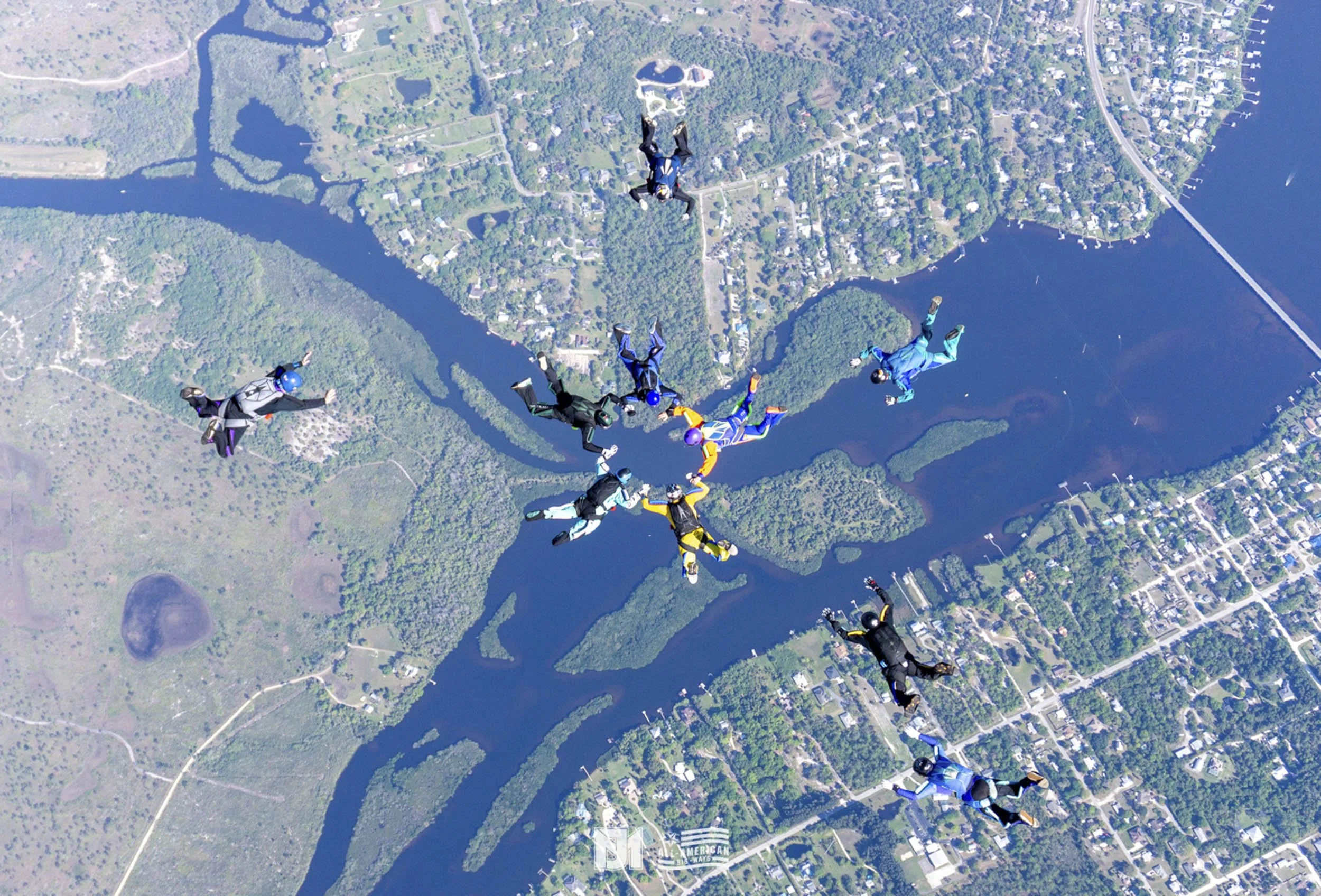 Group of skydivers forming a circle while free-falling over a landscape of lakes, forests, and a residential area.