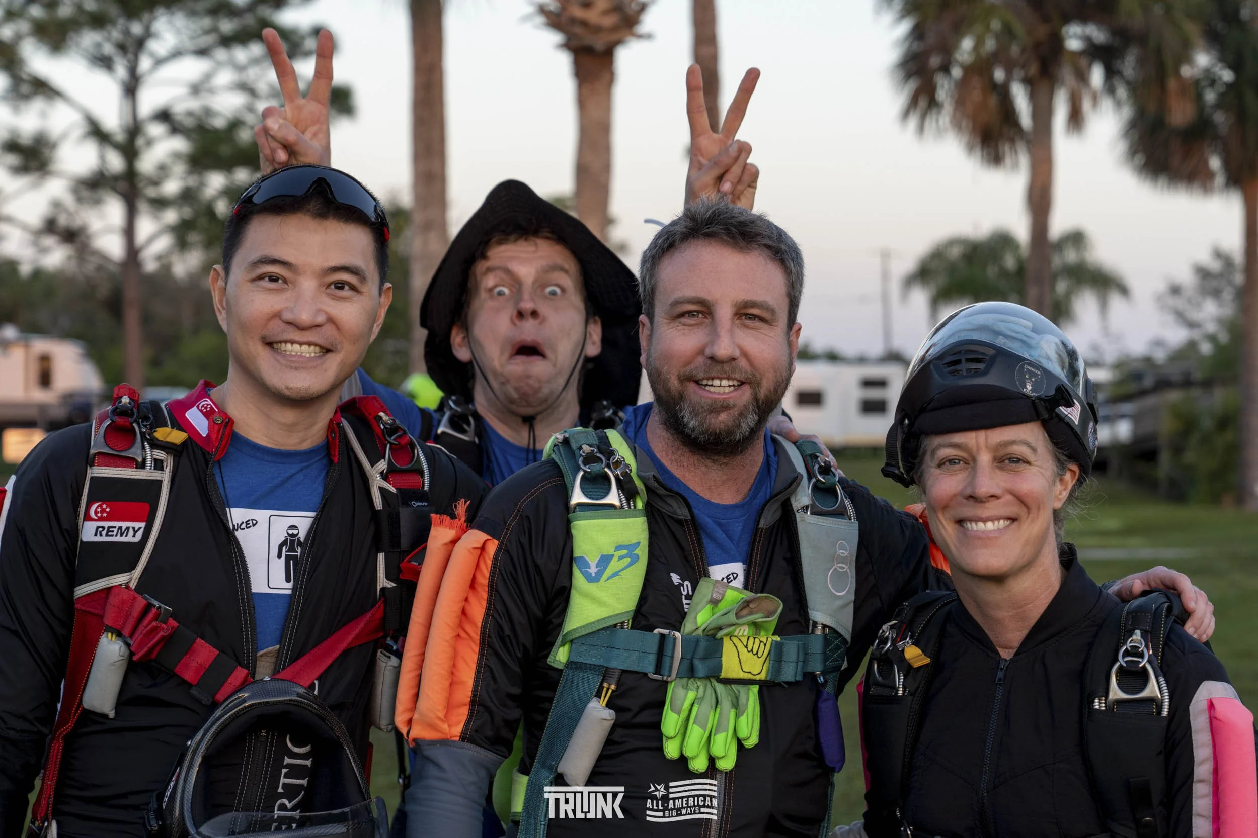 Group of four skydivers smiling and posing for a photo, with trees and a blue sky in the background.