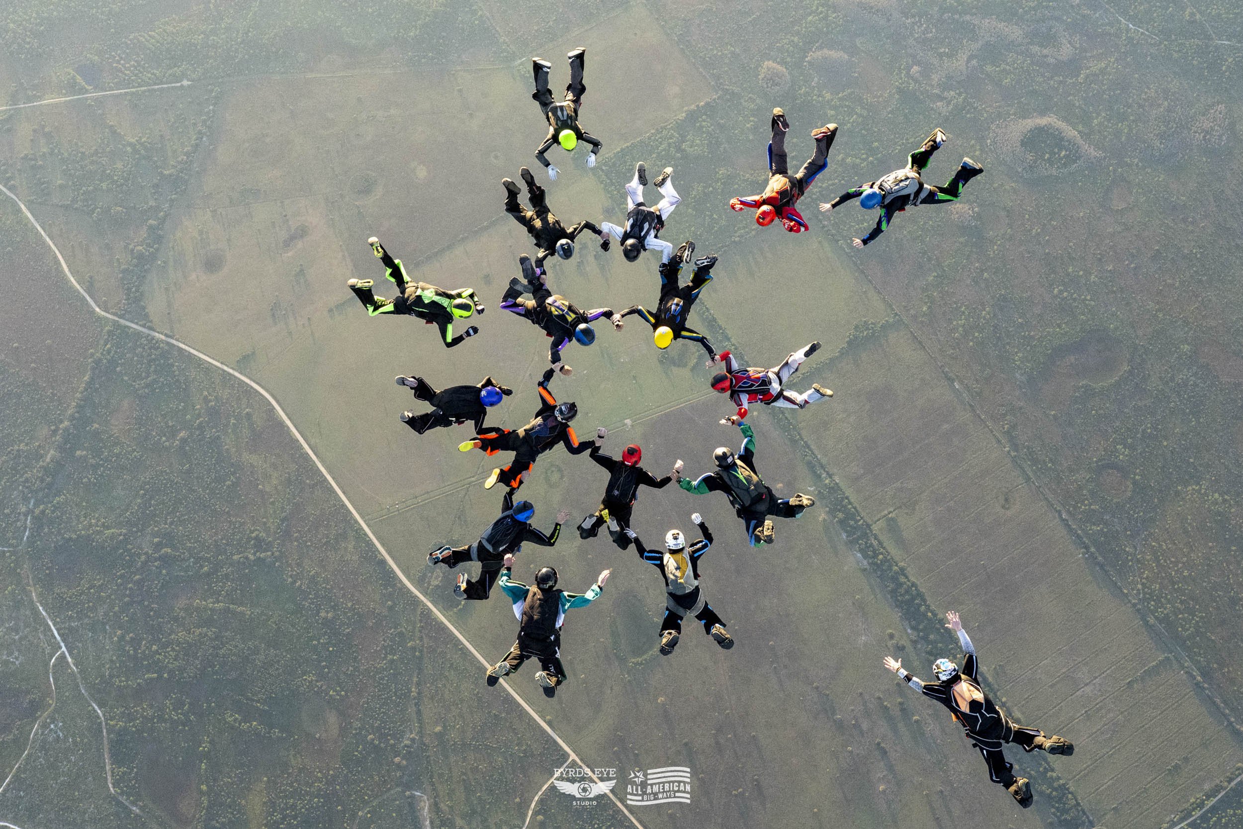 Multiple skydivers in free fall holding hands and forming a circle above a landscape of fields and roads.