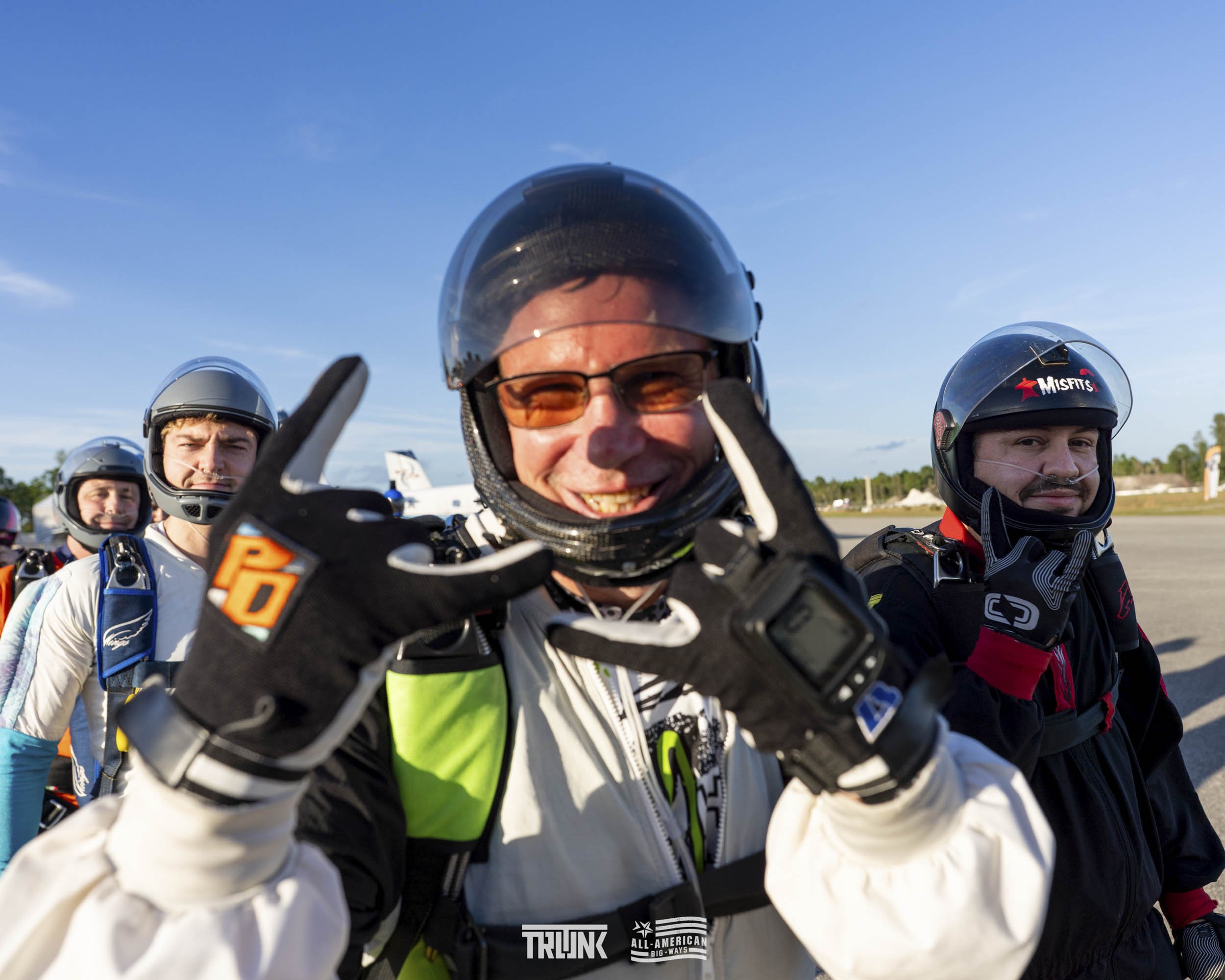 Group of parachutists on the runway wearing helmets and jumpsuits, with the main person smiling and making a hand sign, under a clear blue sky.