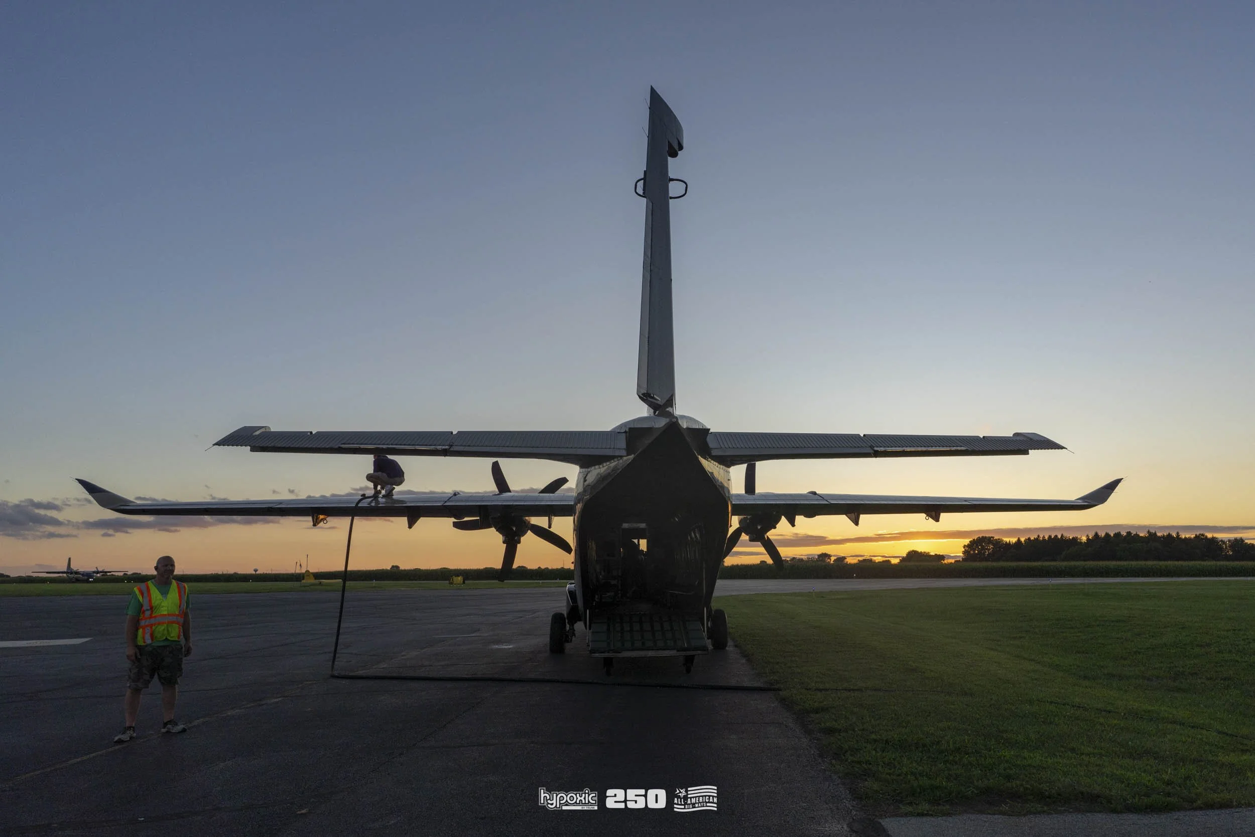 A small aircraft parked on the tarmac at sunset, with a man in a safety vest standing nearby and a person on the wing.