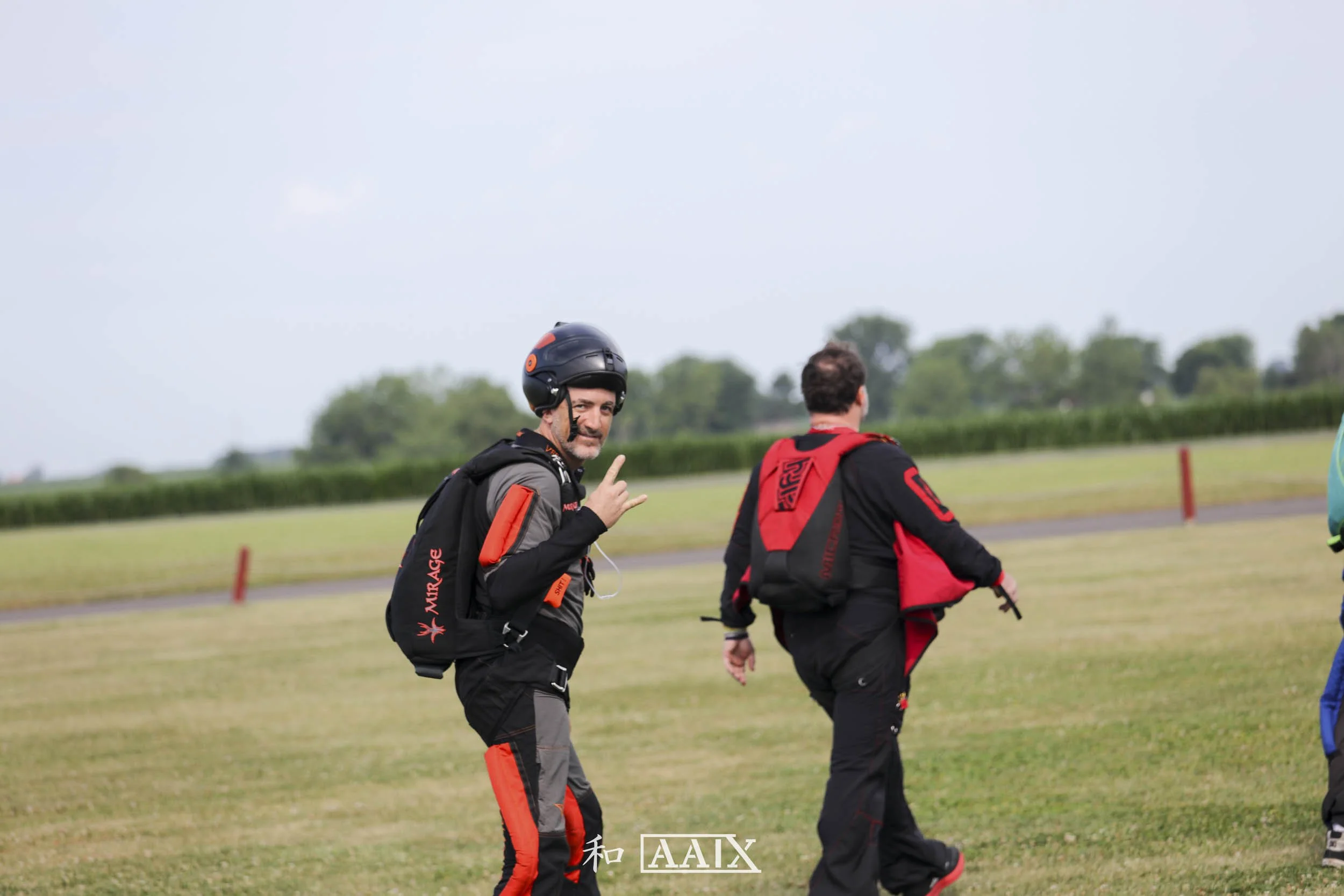 A man wearing a black helmet, black and gray jumpsuit, and a black backpack showing a peace sign and smiling while standing on a grassy field with a cloudy sky in the background.