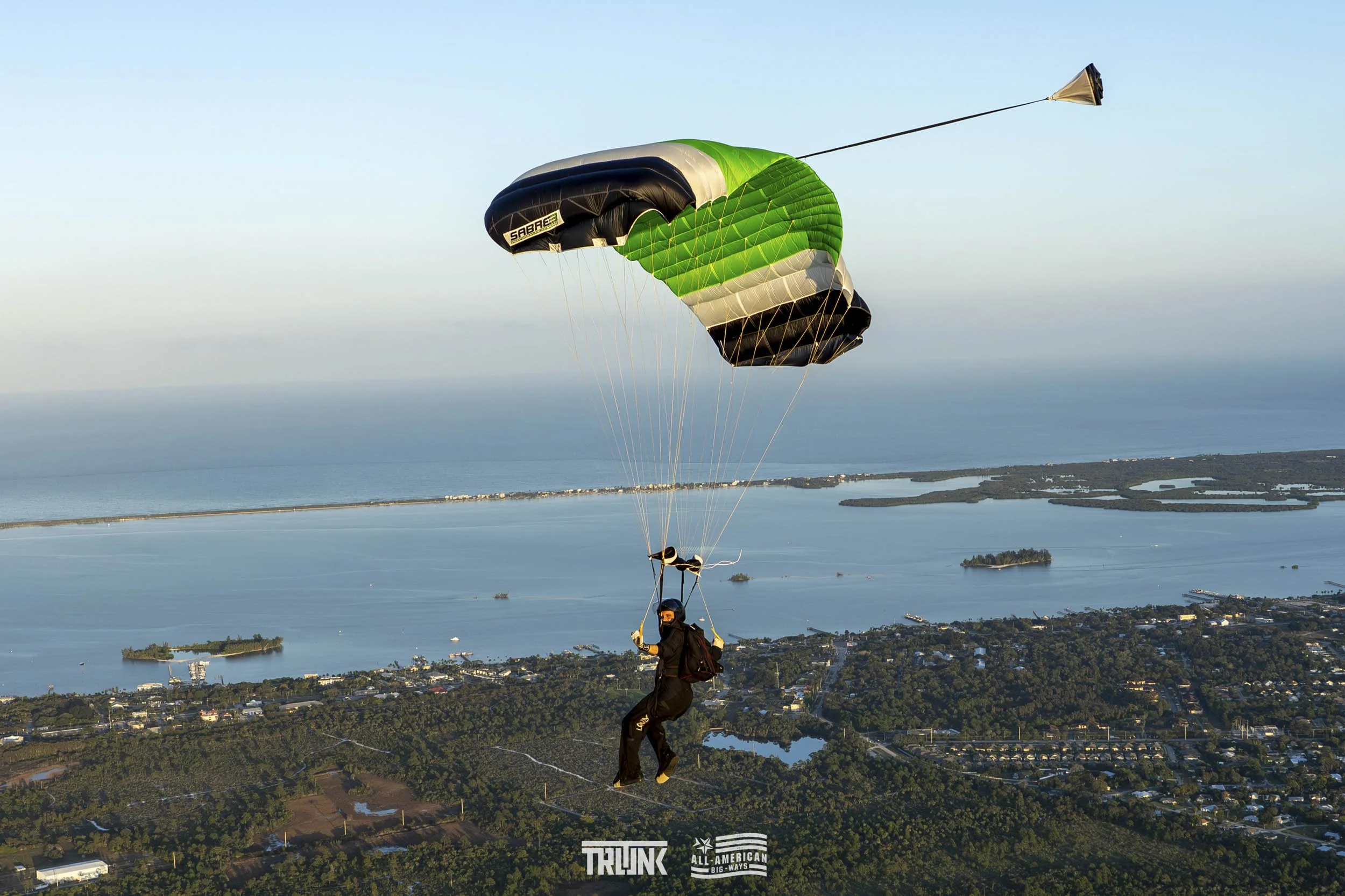 A person skydiving with a black and green parachute over a body of water and landscape.