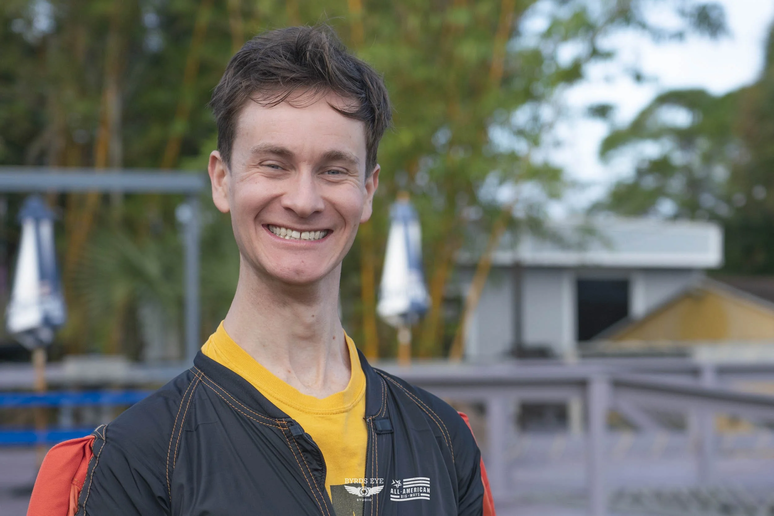 Smiling young man with short brown hair wearing a yellow shirt and a black jacket outdoors.