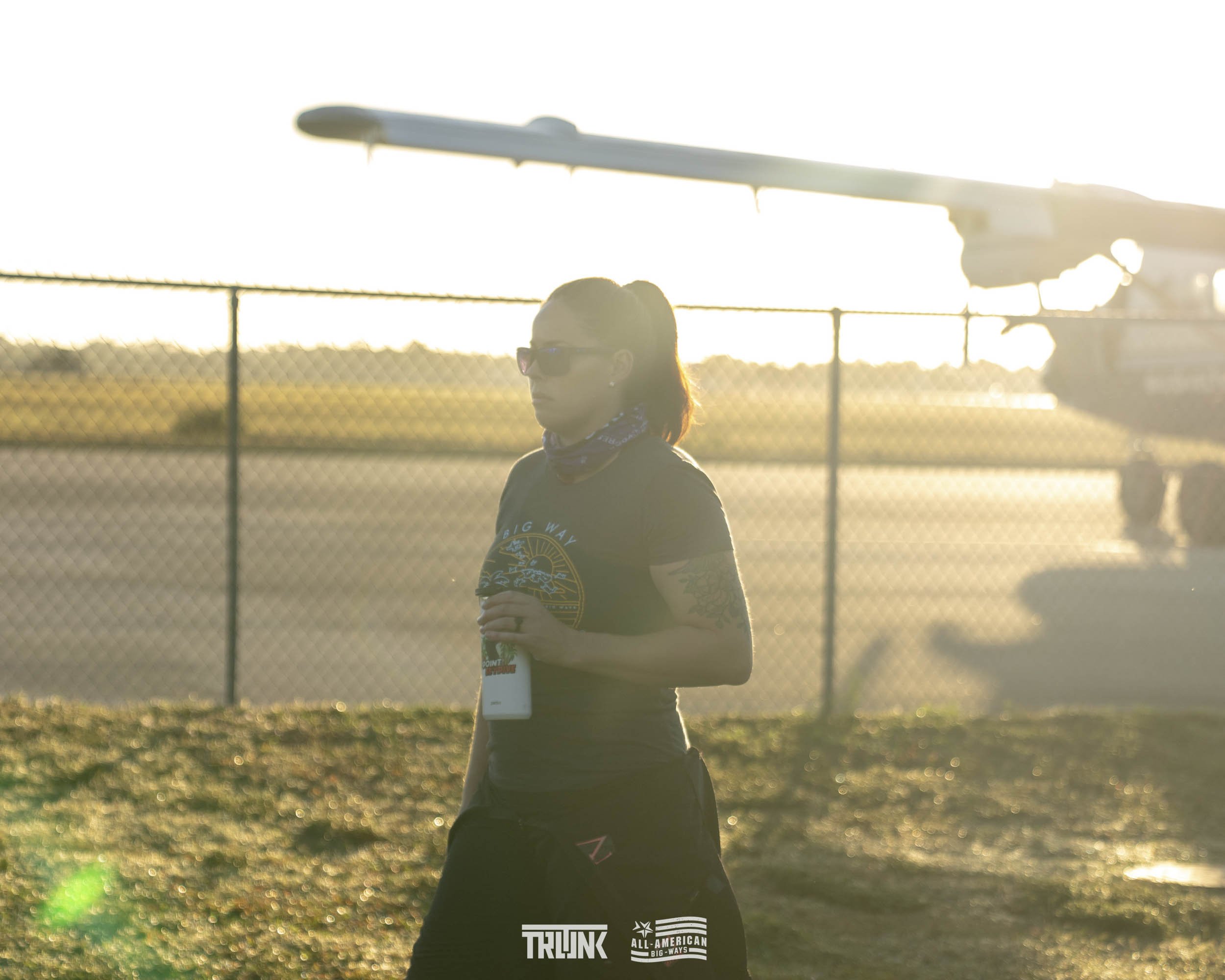 A woman with tattoos and sunglasses walking past an aircraft on a runway with the sun setting in the background.