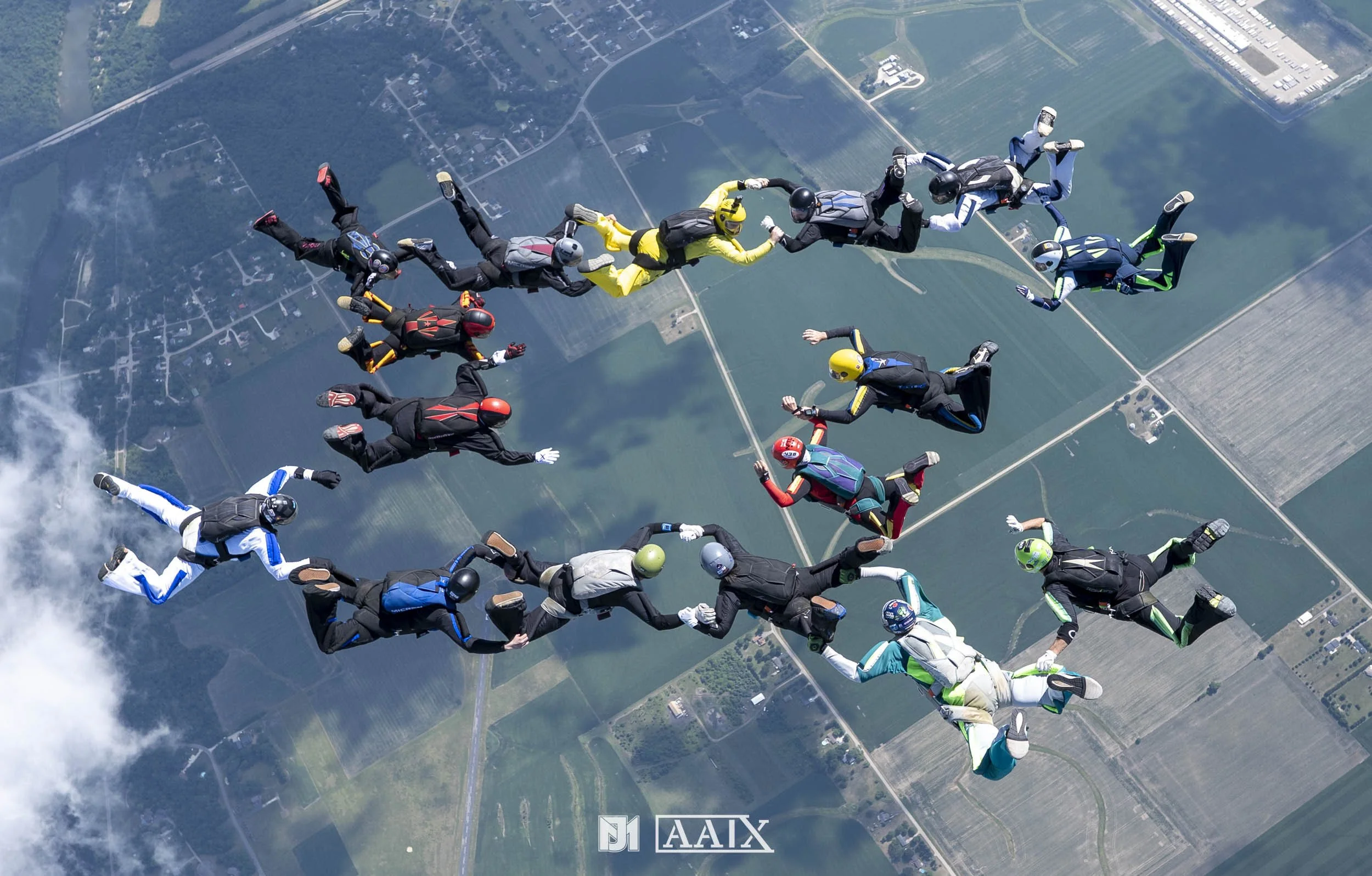 Group of skydivers holding hands in a circle during freefall over farmland.