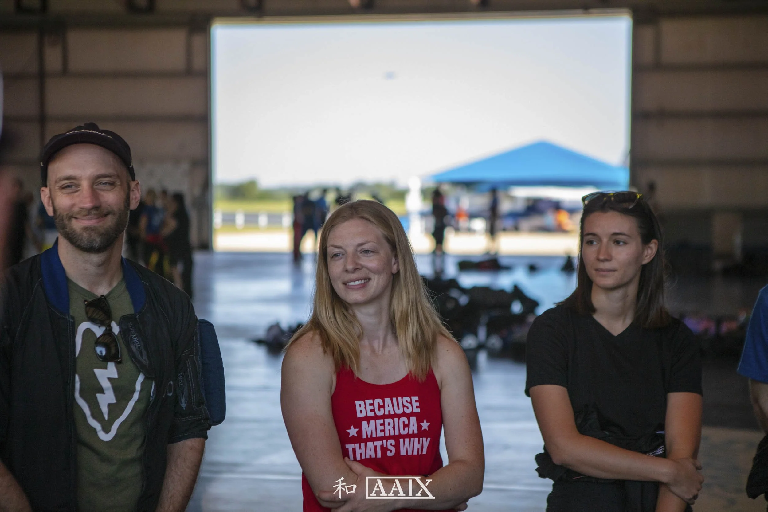 Three people standing together indoors, with a large open door showing a sunny outdoor scene with a blue canopy and people in the background.