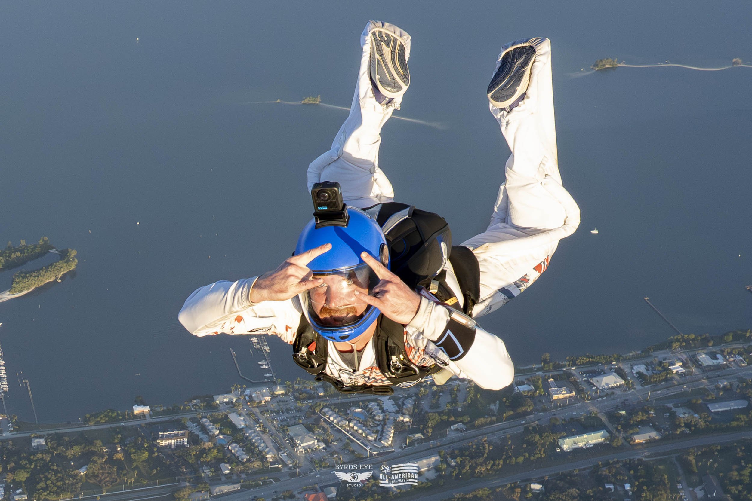 A skydiver in a white jumpsuit with black and orange designs, wearing a blue helmet and goggles, free-falling above a body of water and a cityscape below.