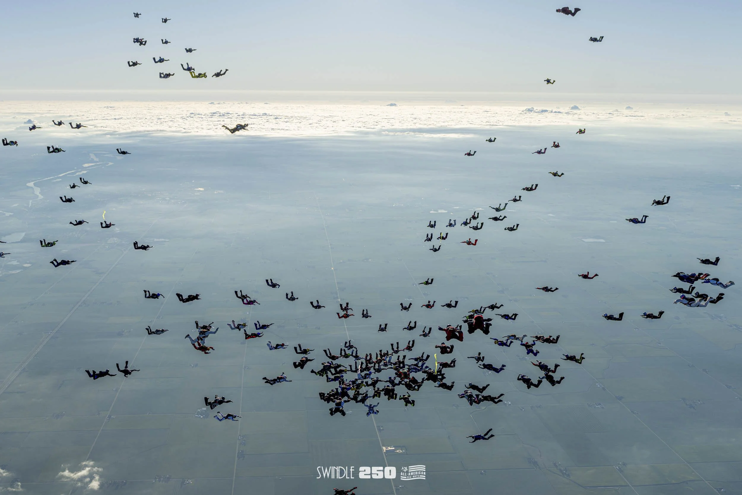 Parachutists in colorful jumpsuits descending over a snowy landscape during an event, with a logo that says 'SWINDLE 250' at the bottom.