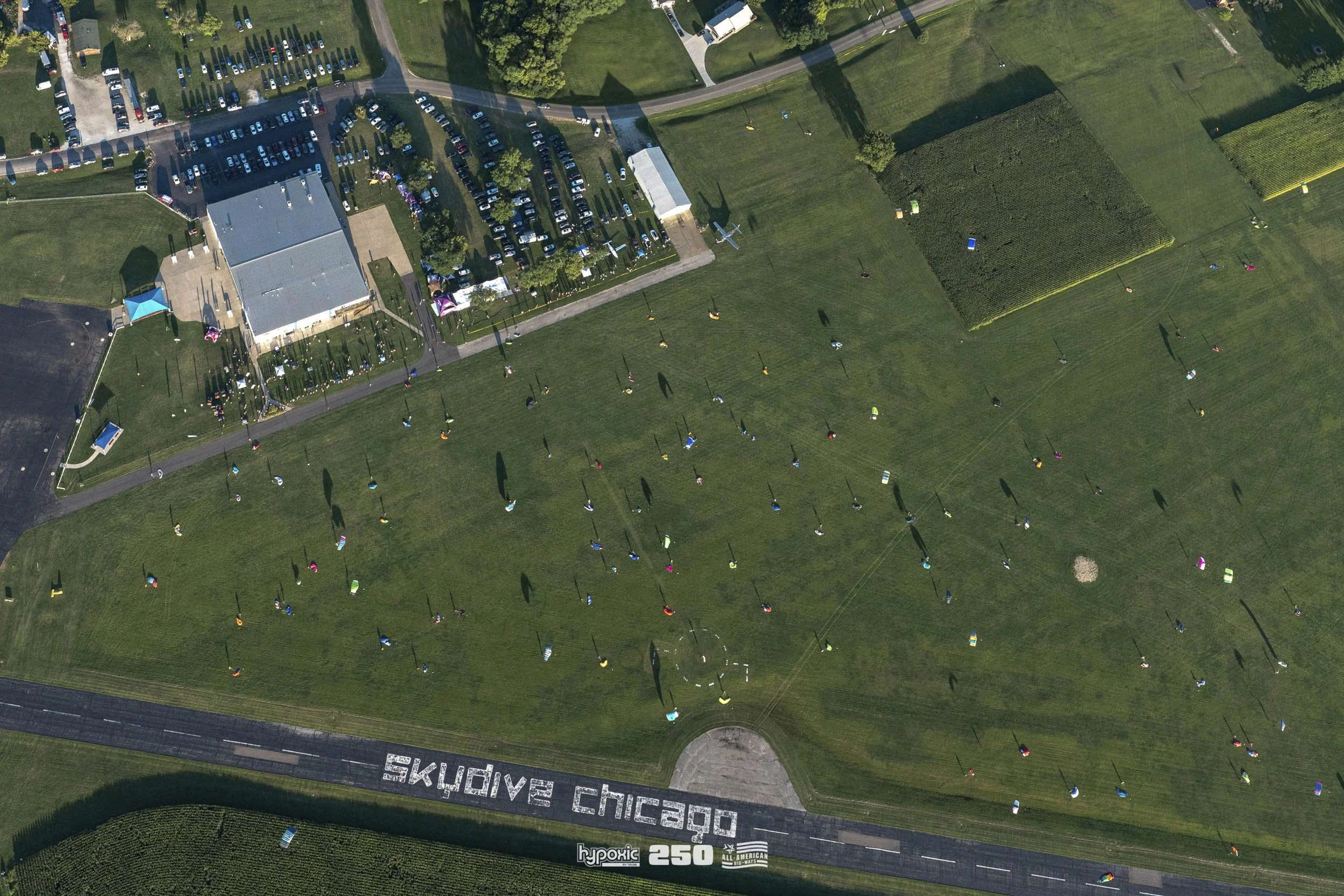 Aerial view of a park with various colorful wind sculptures scattered across the grassy field. A building with parking spaces and cars is visible at the top left, and a large message spelled out on the ground reads 'EXPLORE CHICAGO.'