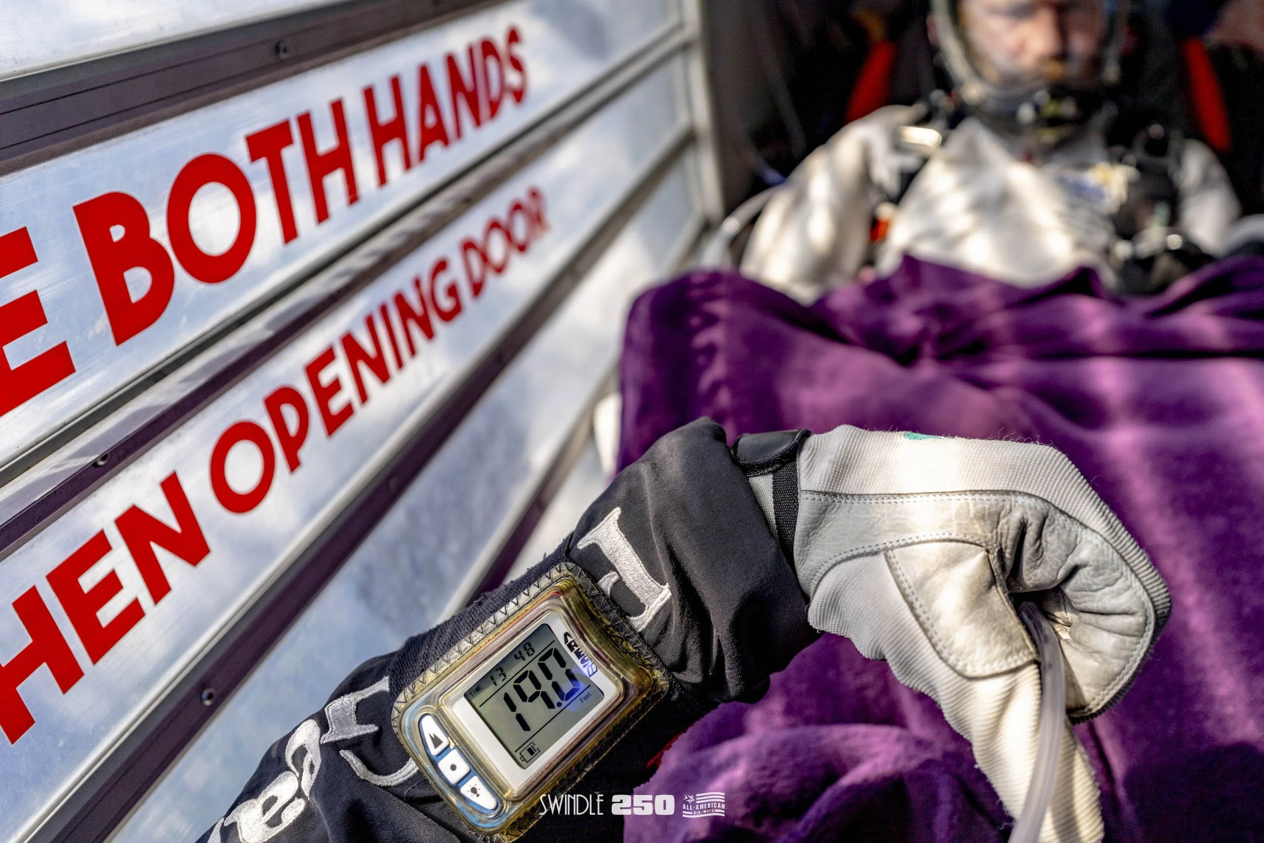 Close-up of a person's gloved forearm and wristwatch, with the person lying on a purple blanket inside an ambulance. The person's face is blurred in the background. A sign with red letters reading 'THE BOTH HANDS' and other words is partially visible