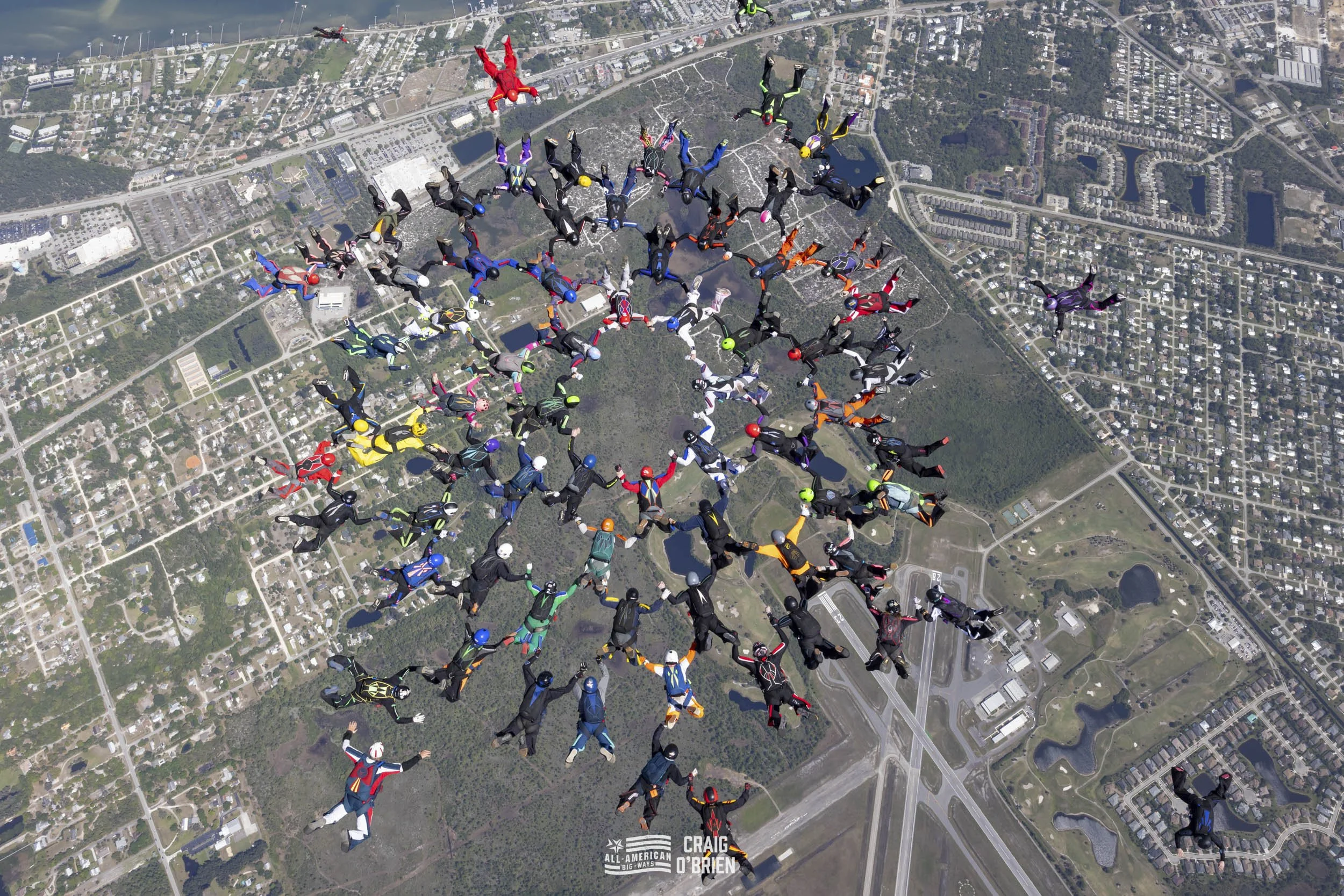 A large group of skydivers in colorful jumpsuits and helmets free falling in formation over a city landscape with roads, buildings, and green areas.