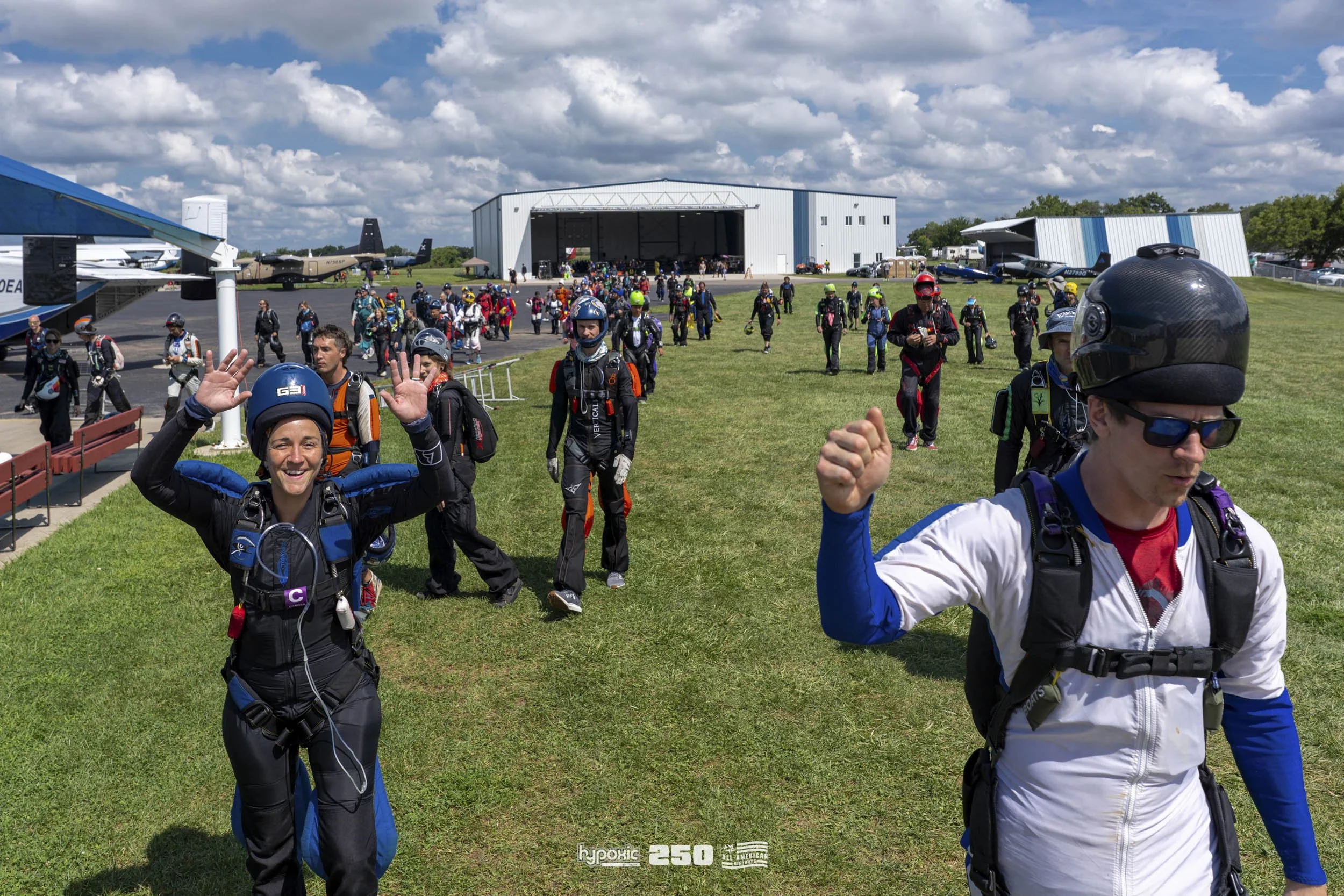 A group of skydivers on the grass near hangars and airplanes at a skydiving facility, with some smiling and waving, preparing for a jump on a partly cloudy day.