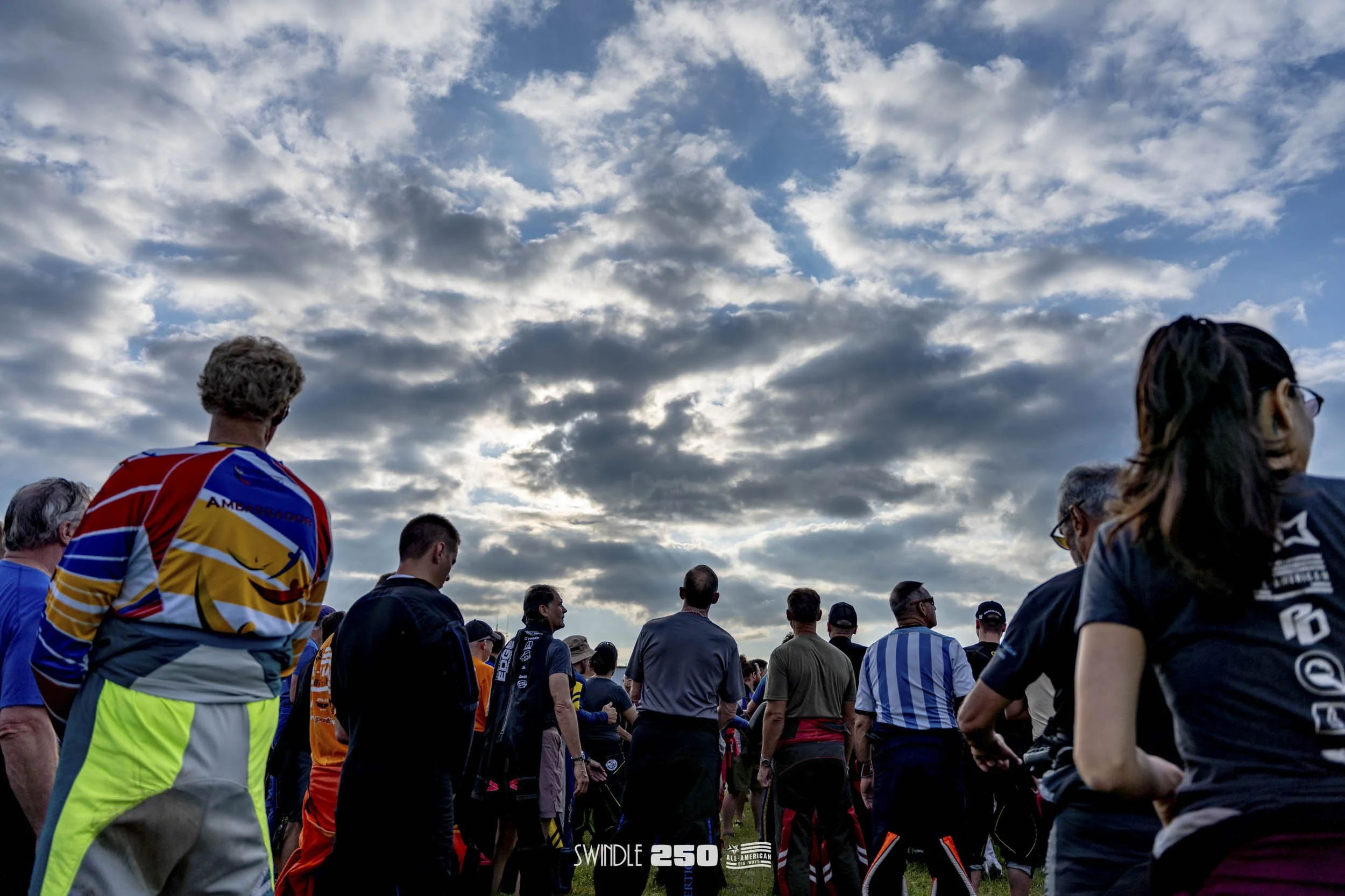 A crowd of people gathered outdoors during daytime, facing away from the camera, under a cloudy sky.