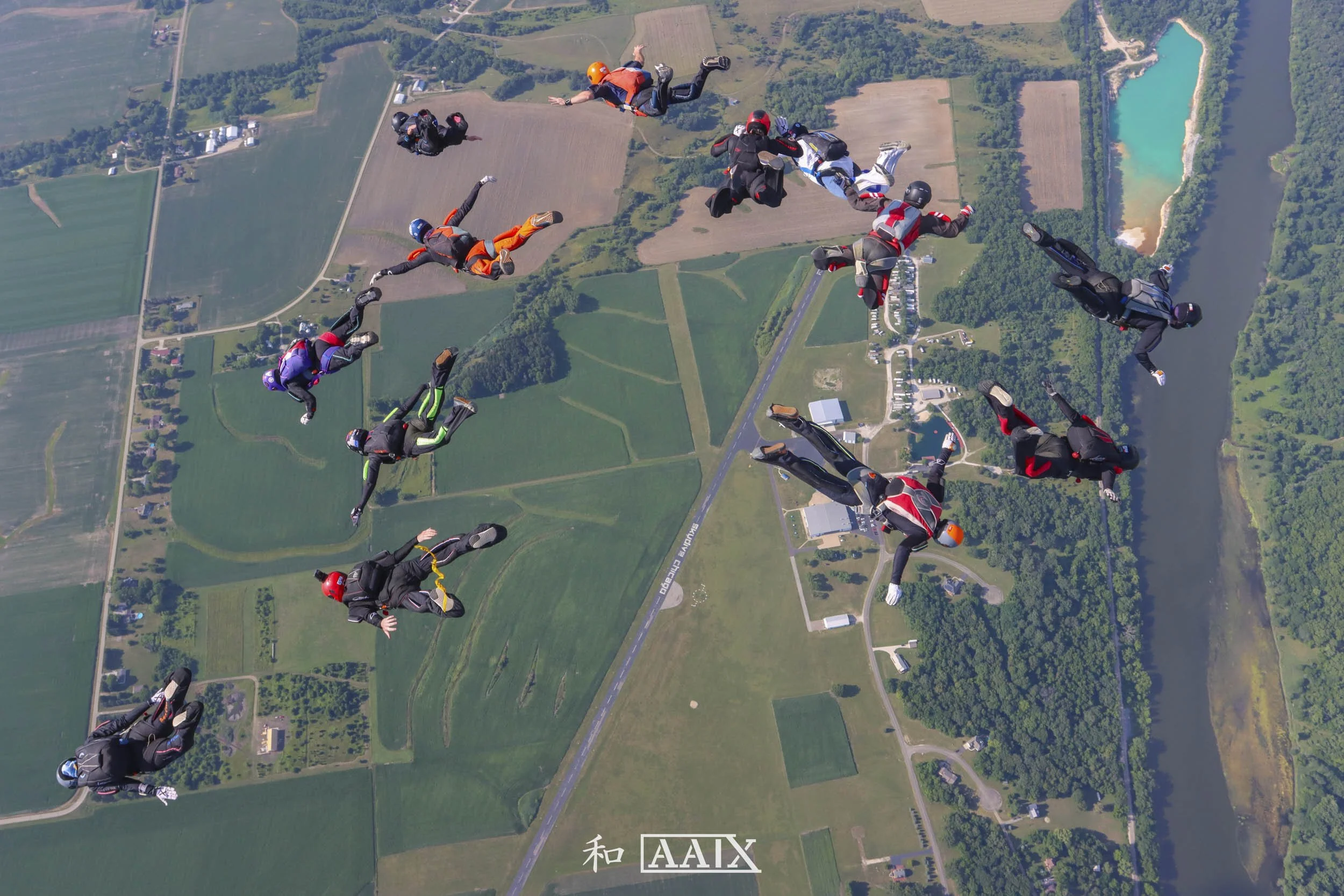 A group of skydivers in free fall over a rural landscape with fields, trees, a river, and a small town.