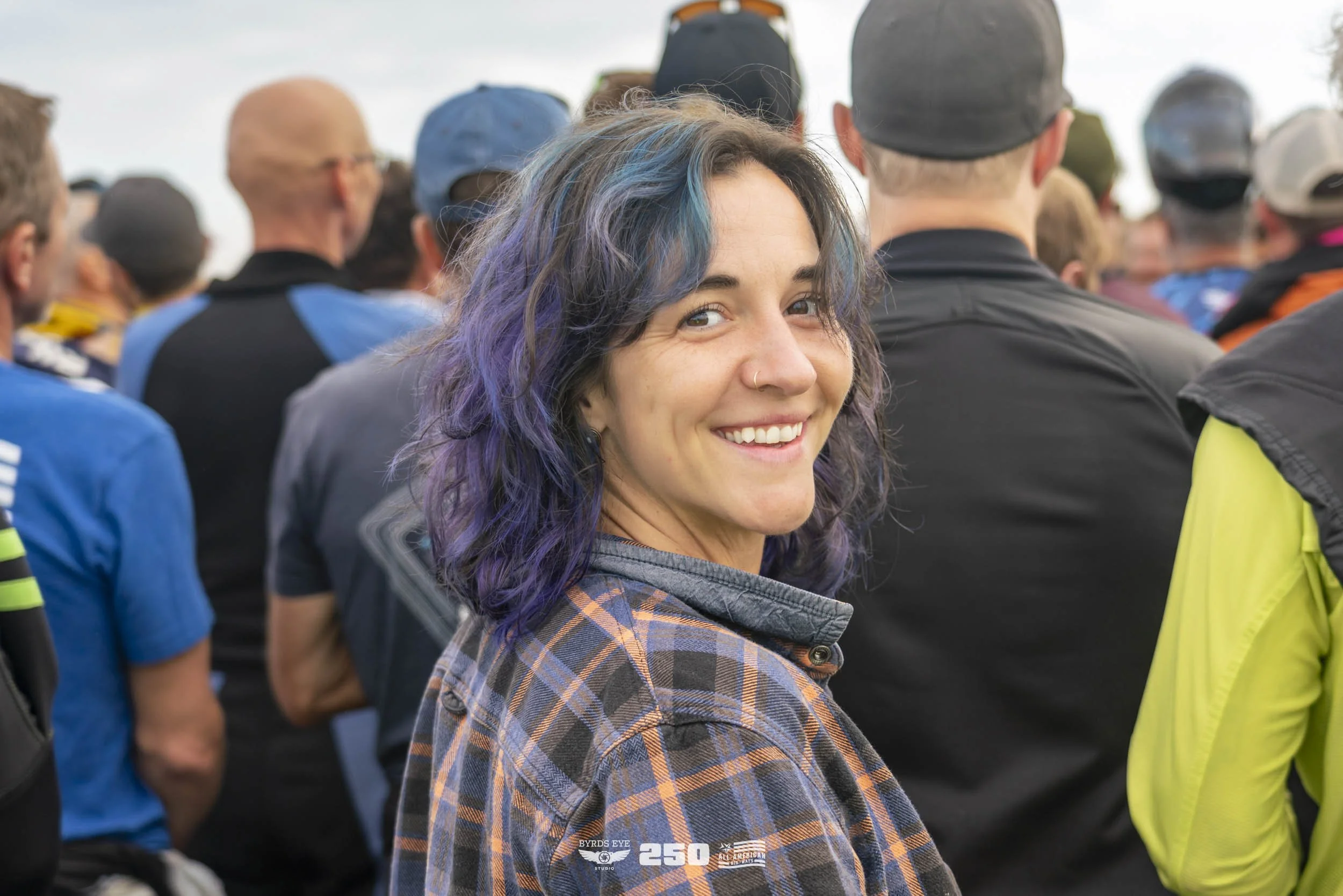 Young woman with purple and blue hair smiling at crowd gathering outdoors.