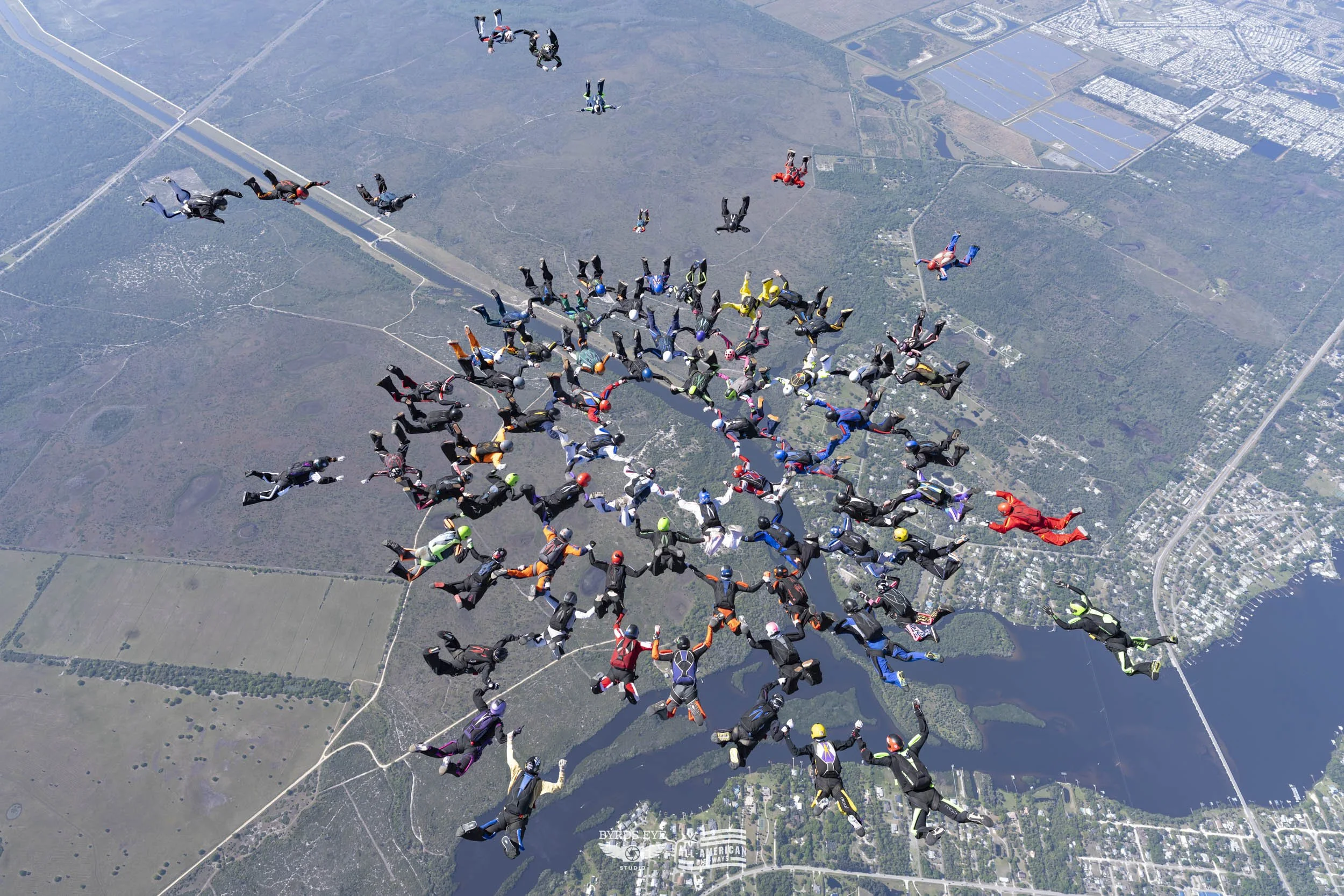 Group of skydivers in freefall formation over a landscape with lakes and urban areas.