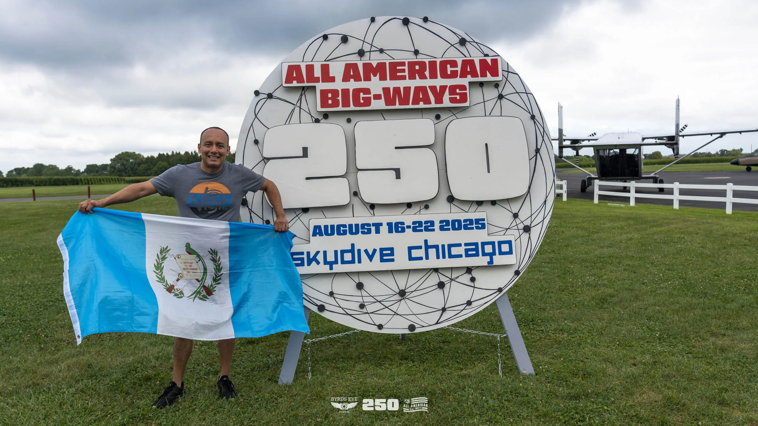 A man smiling and holding a Guatemala flag stands next to a large sign that reads "All American Big-Ways 250" with the event date "August 16-22 2025" at Skydive Chicago. There is a small aircraft and a runway in the background under a cloudy sky.