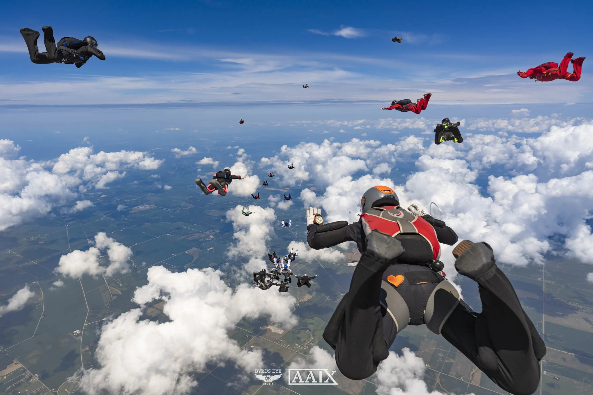 A group of skydivers in freefall above the clouds and landscape below during daytime, with some skydivers wearing colorful suits and helmets.