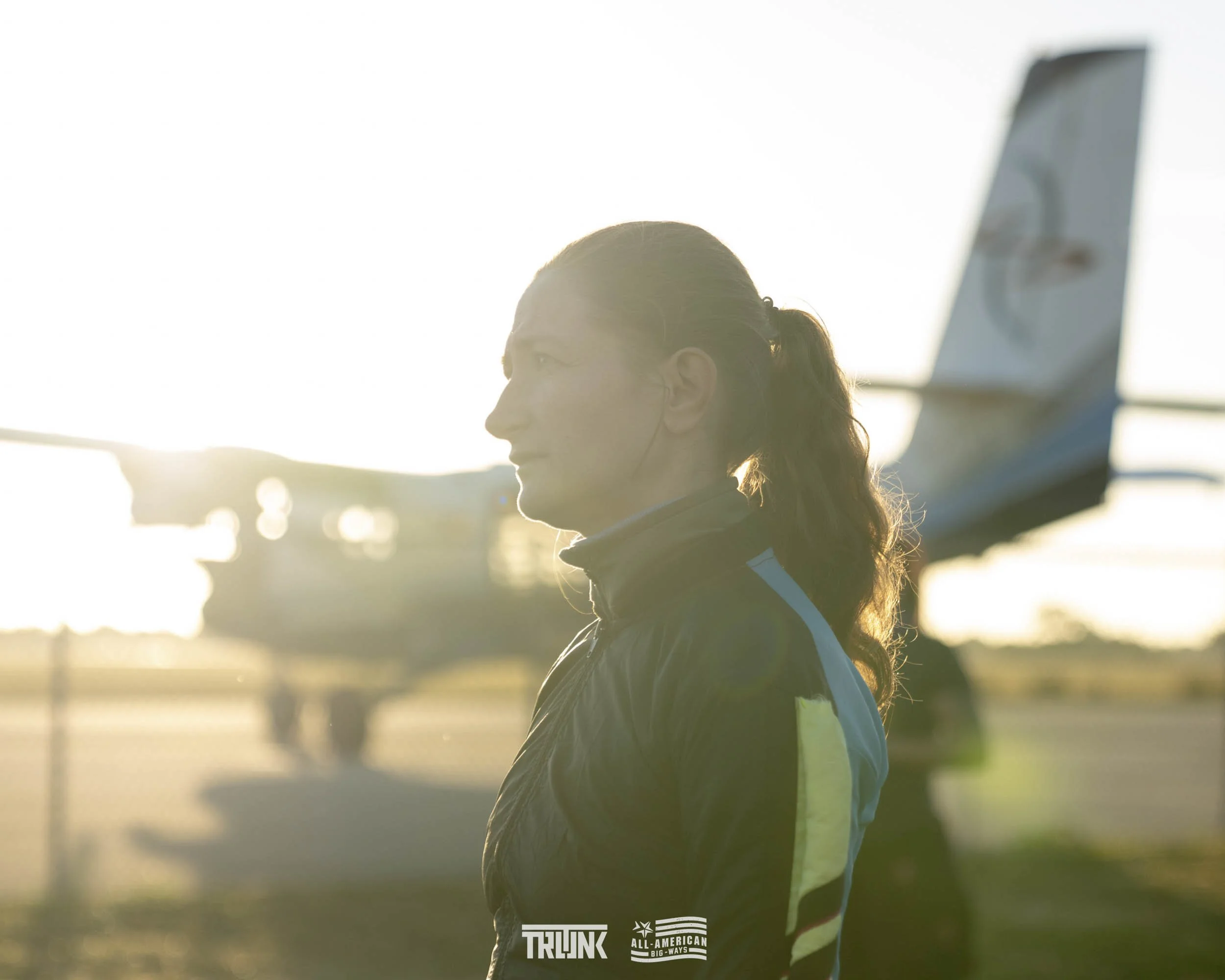 A woman standing outdoors near an airplane at sunrise or sunset, with her profile visible and the airplane behind her.
