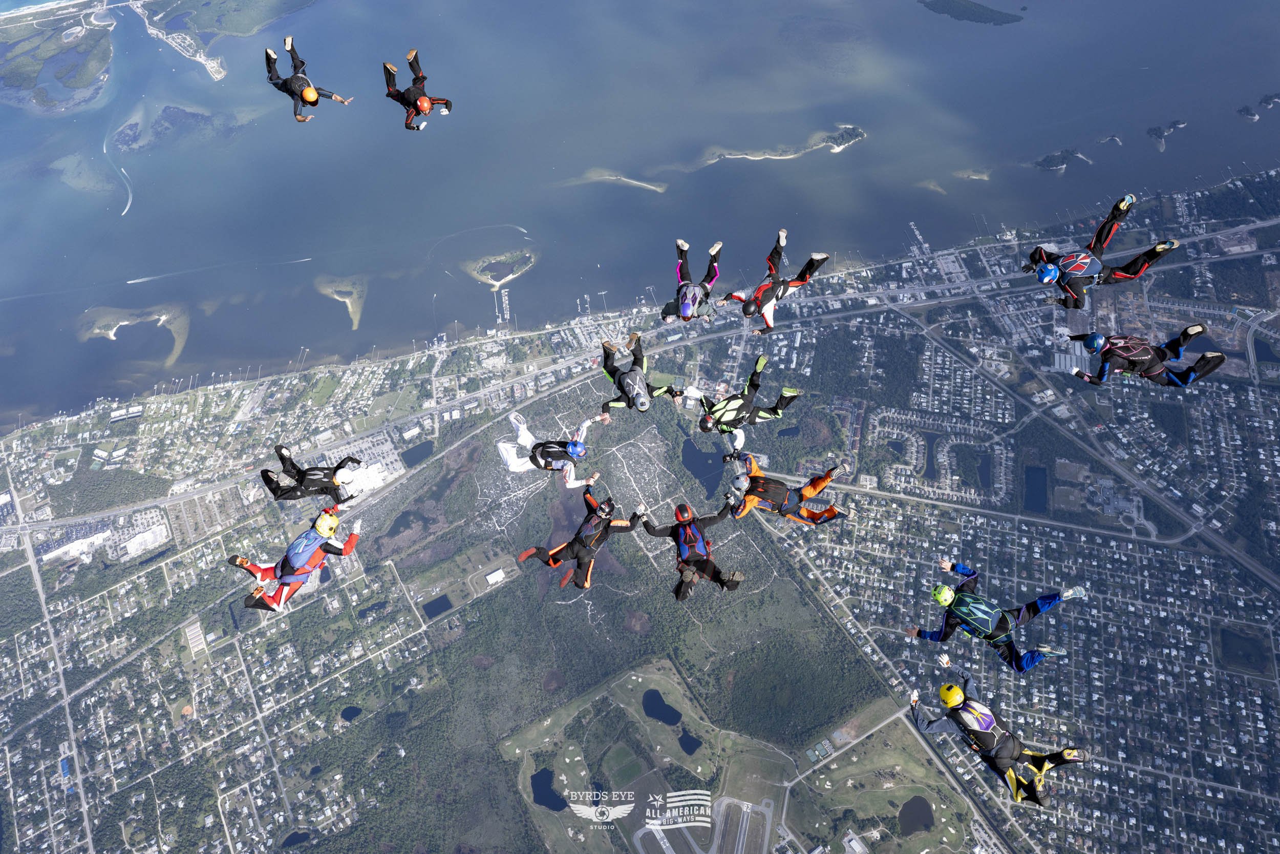 Group of skydivers in freefall over a city and lake, holding hands in a circle and wearing colorful jumpsuits and helmets.