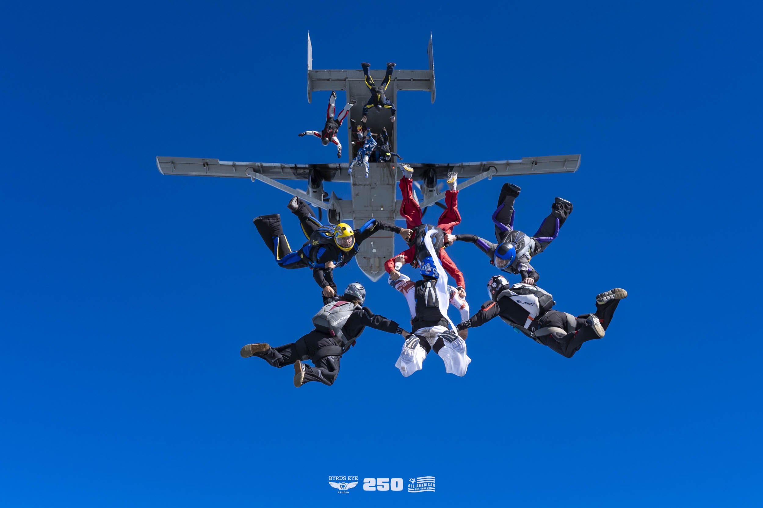 A group of skydivers performs a coordinated stunt in freefall against a clear blue sky, with one skydiver hanging upside down from an aircraft.