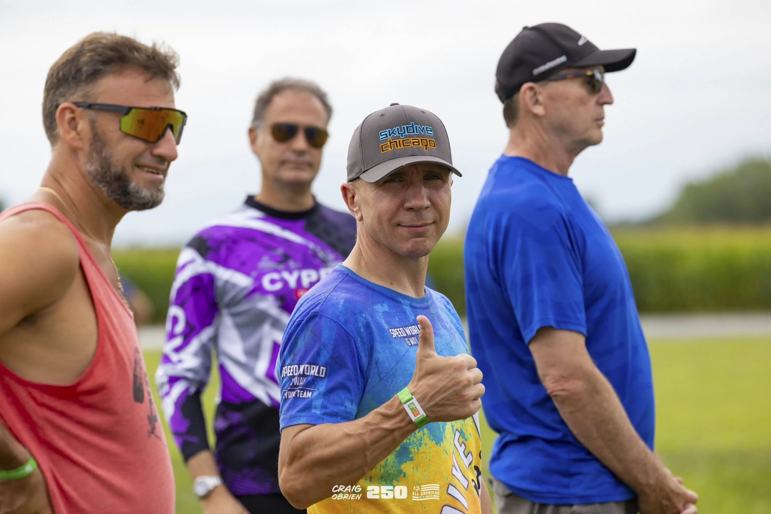 Group of four men standing outdoors in athletic attire, one giving a thumbs up, with a grassy field and cloudy sky in the background.
