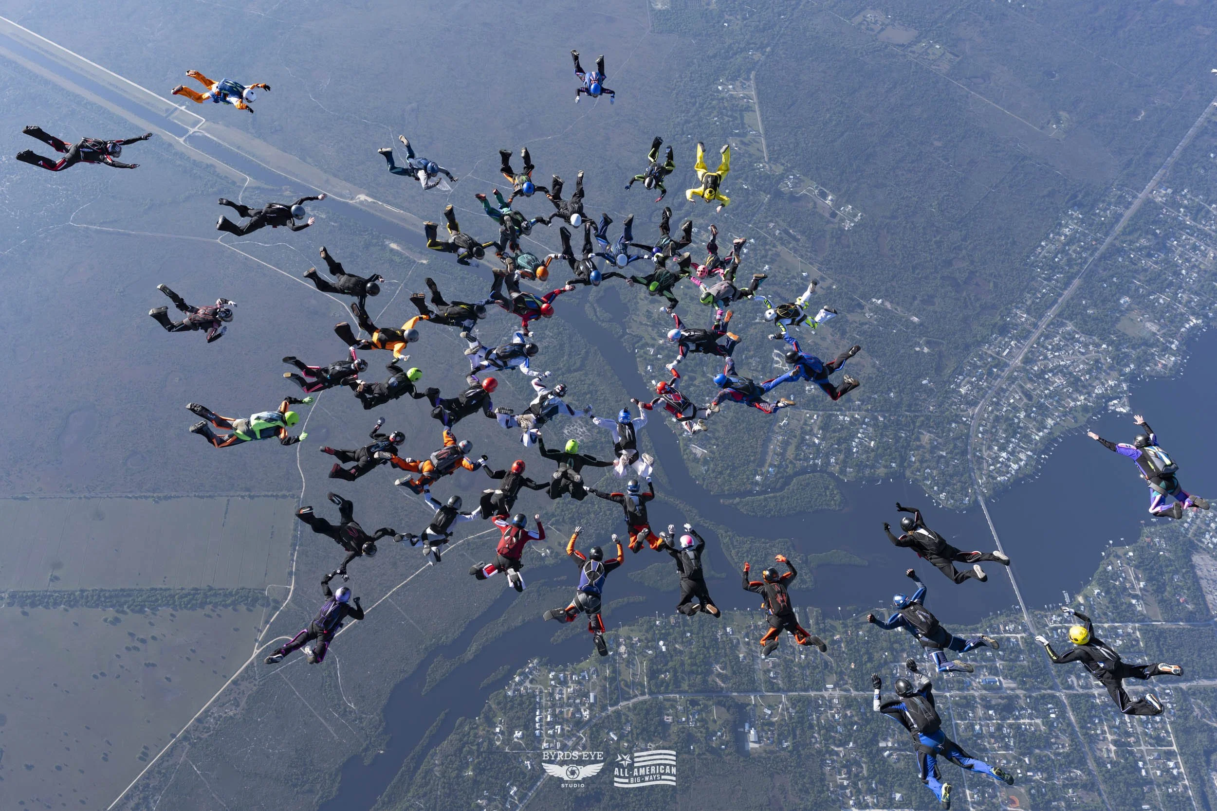 Group of 31 skydivers in freefall formation over a landscape with rivers and a town below.