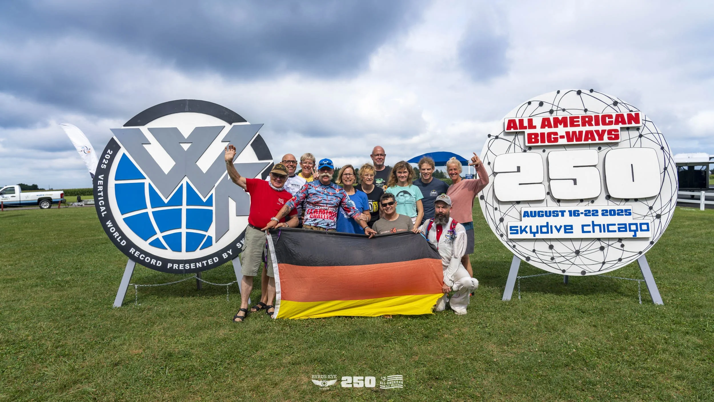 Group of people celebrating at Skydive Chicago for the All American Big-Ways record, holding a German flag, with large signs and logos supporting the event on a grassy field with cloudy sky.